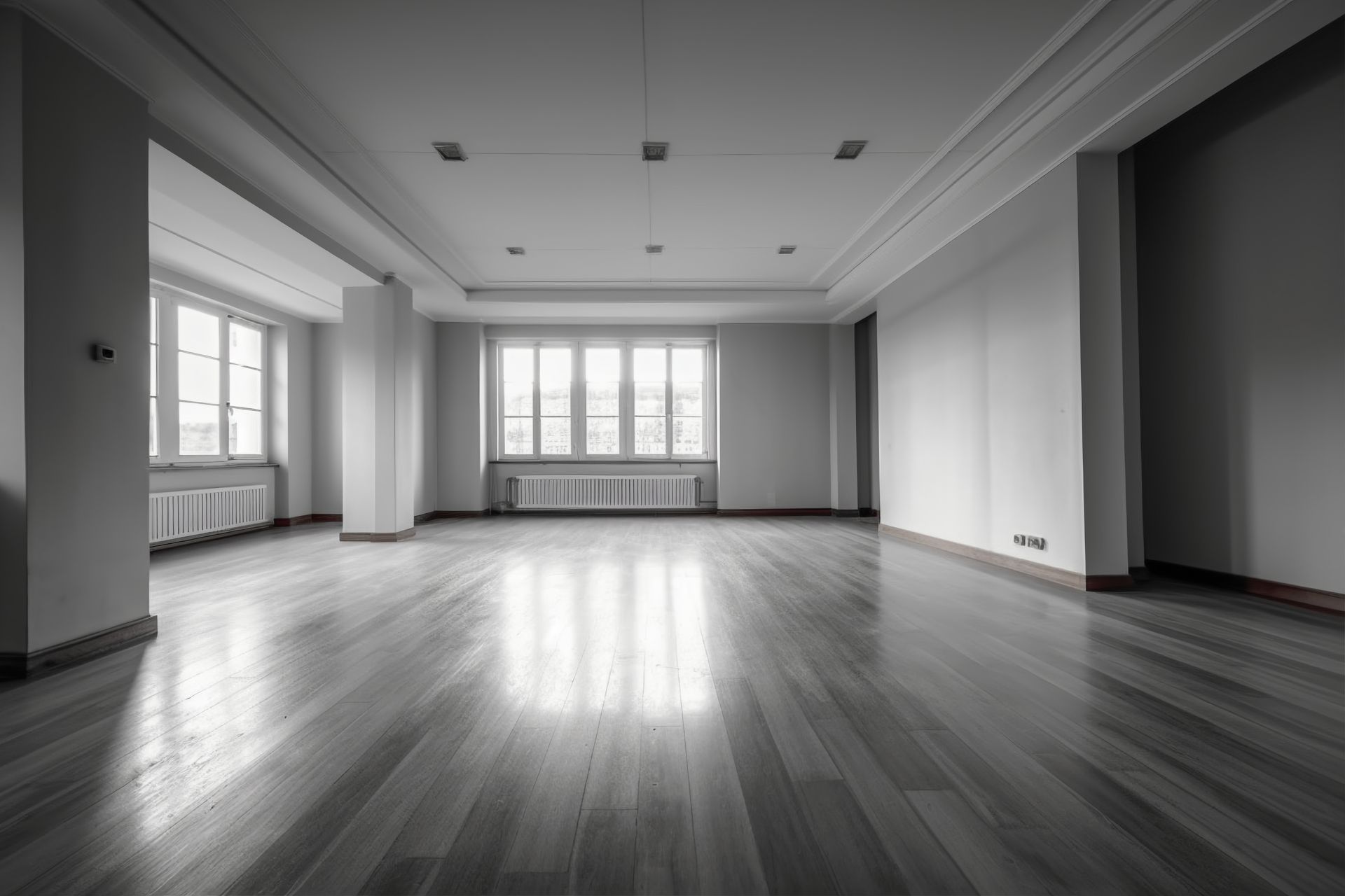 Empty, sunlit room with hardwood floors, large windows, and white walls.