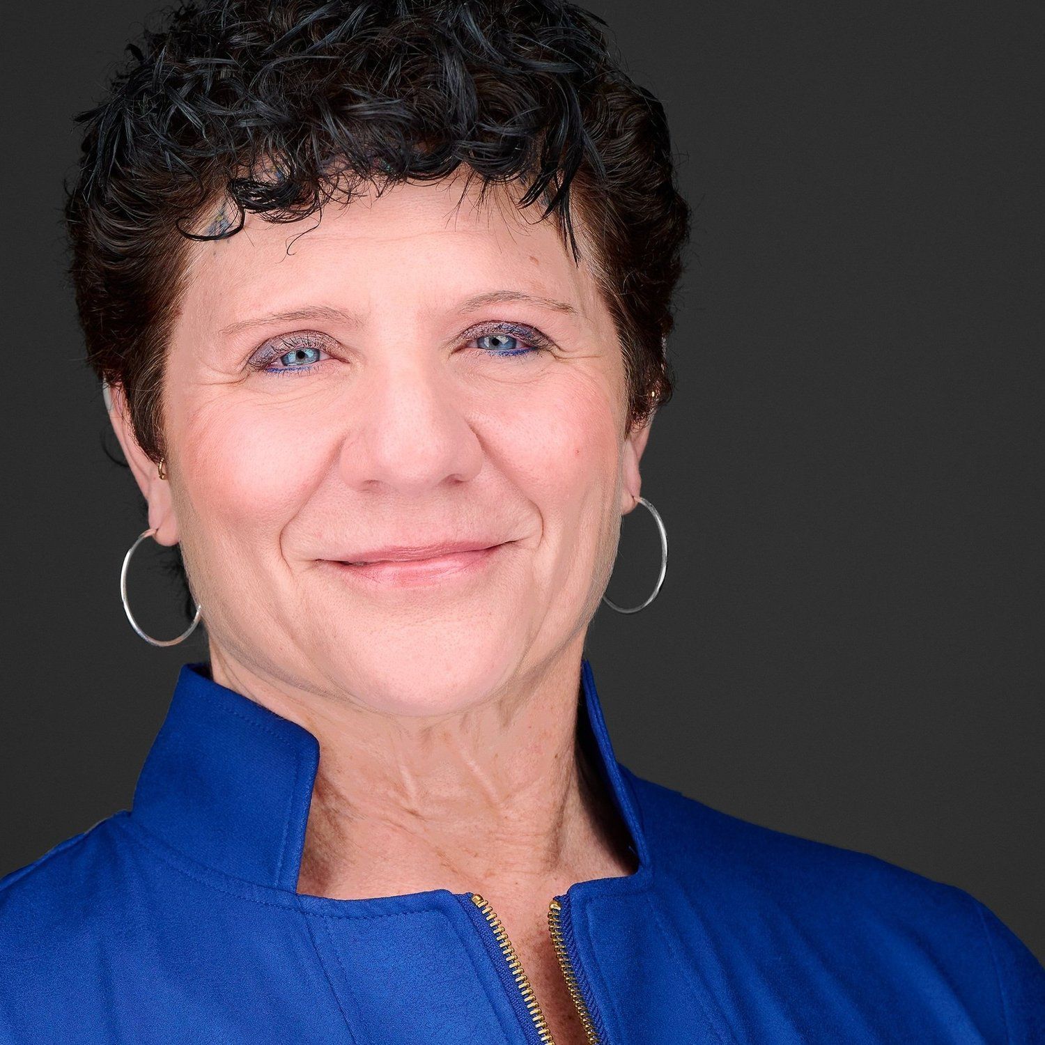 A woman wearing a blue jacket and hoop earrings smiles for the camera