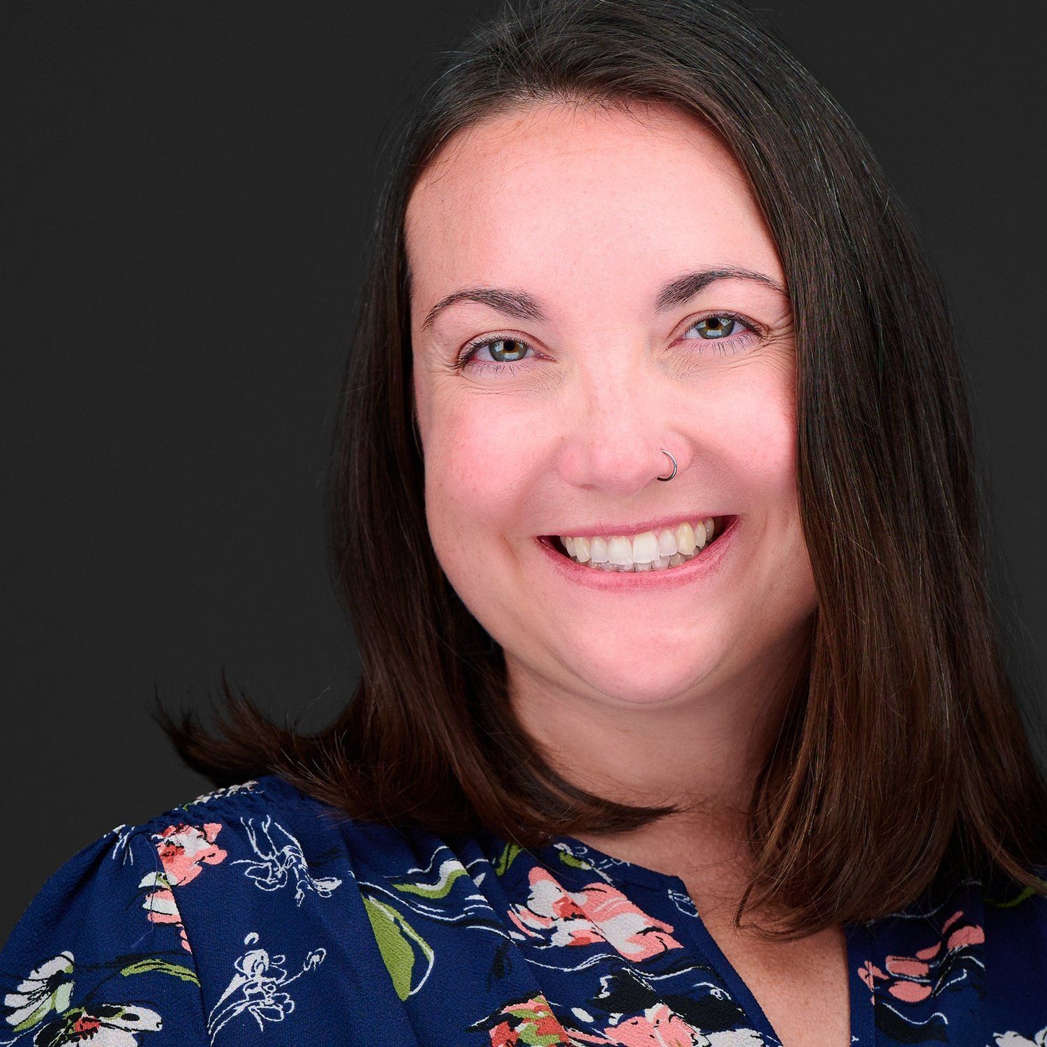 Woman with brown hair and a floral shirt smiles at the camera against a dark background.