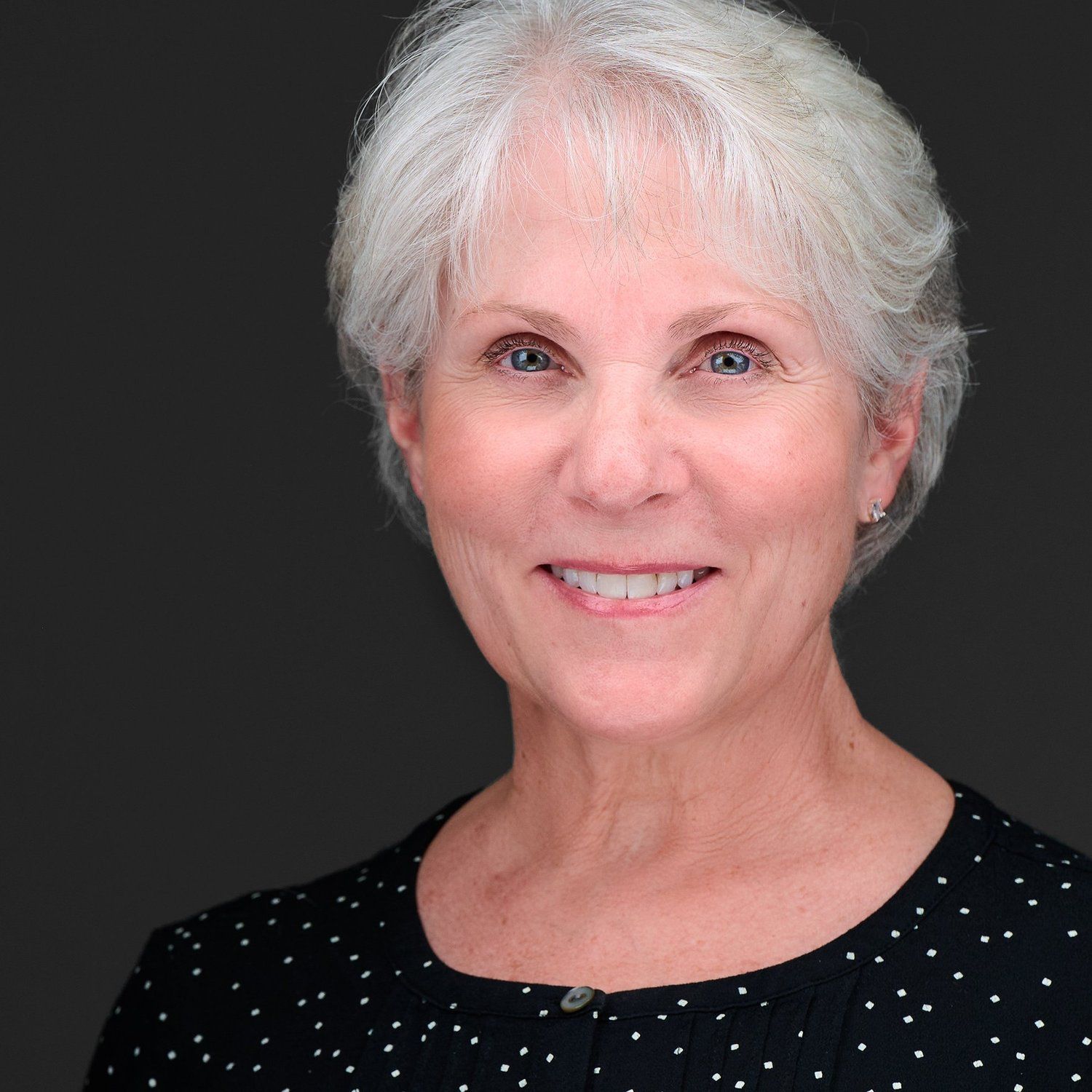 A woman with white hair is smiling and wearing a black and white polka dot shirt.