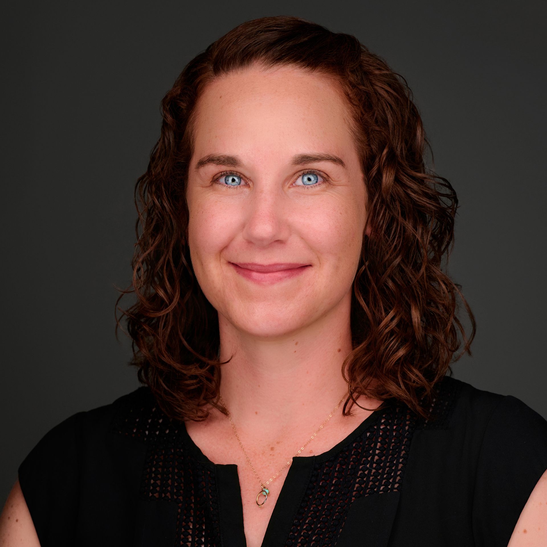 A woman with curly hair and blue eyes is wearing a black shirt and a necklace.