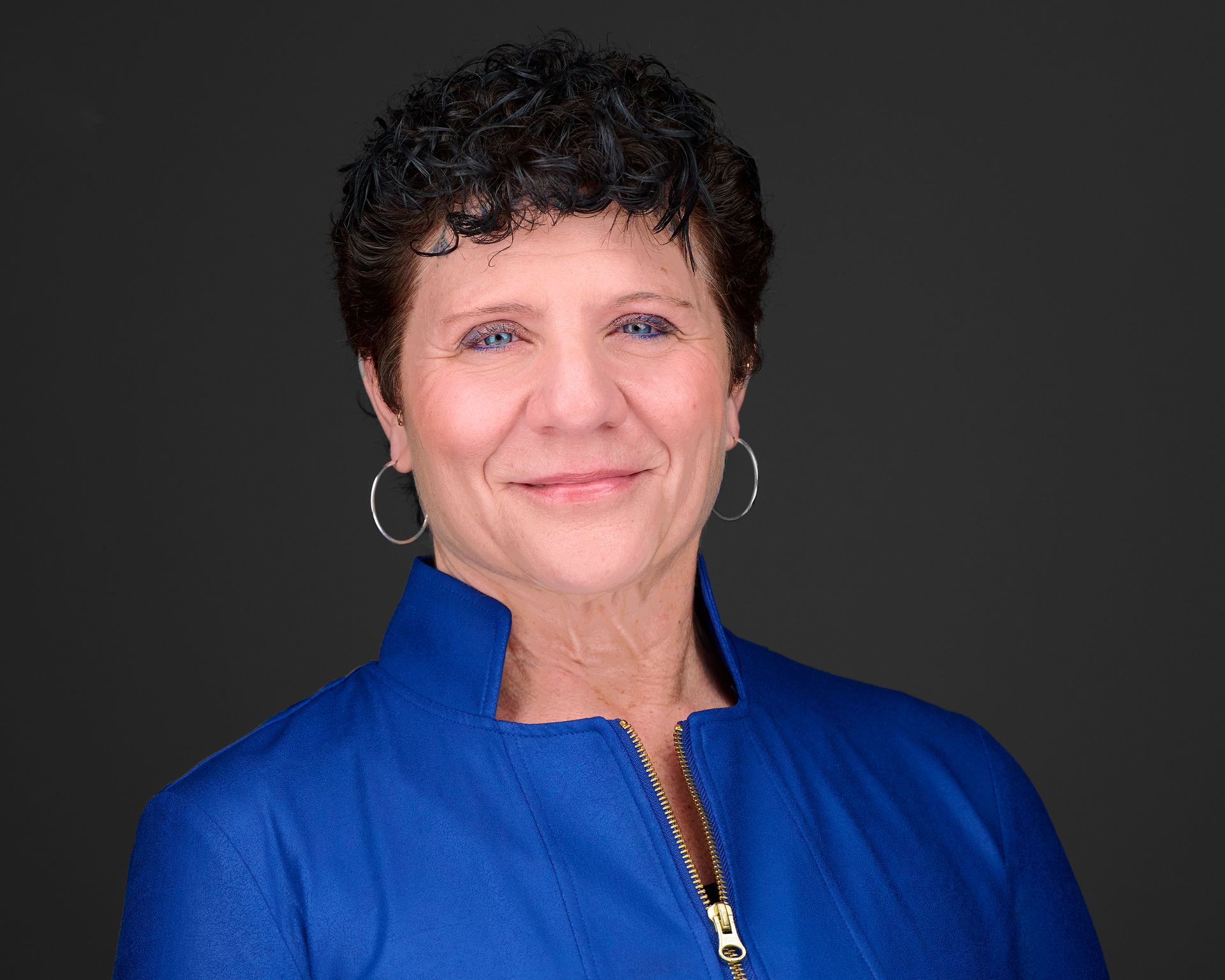 A woman wearing a blue jacket and hoop earrings smiles for the camera