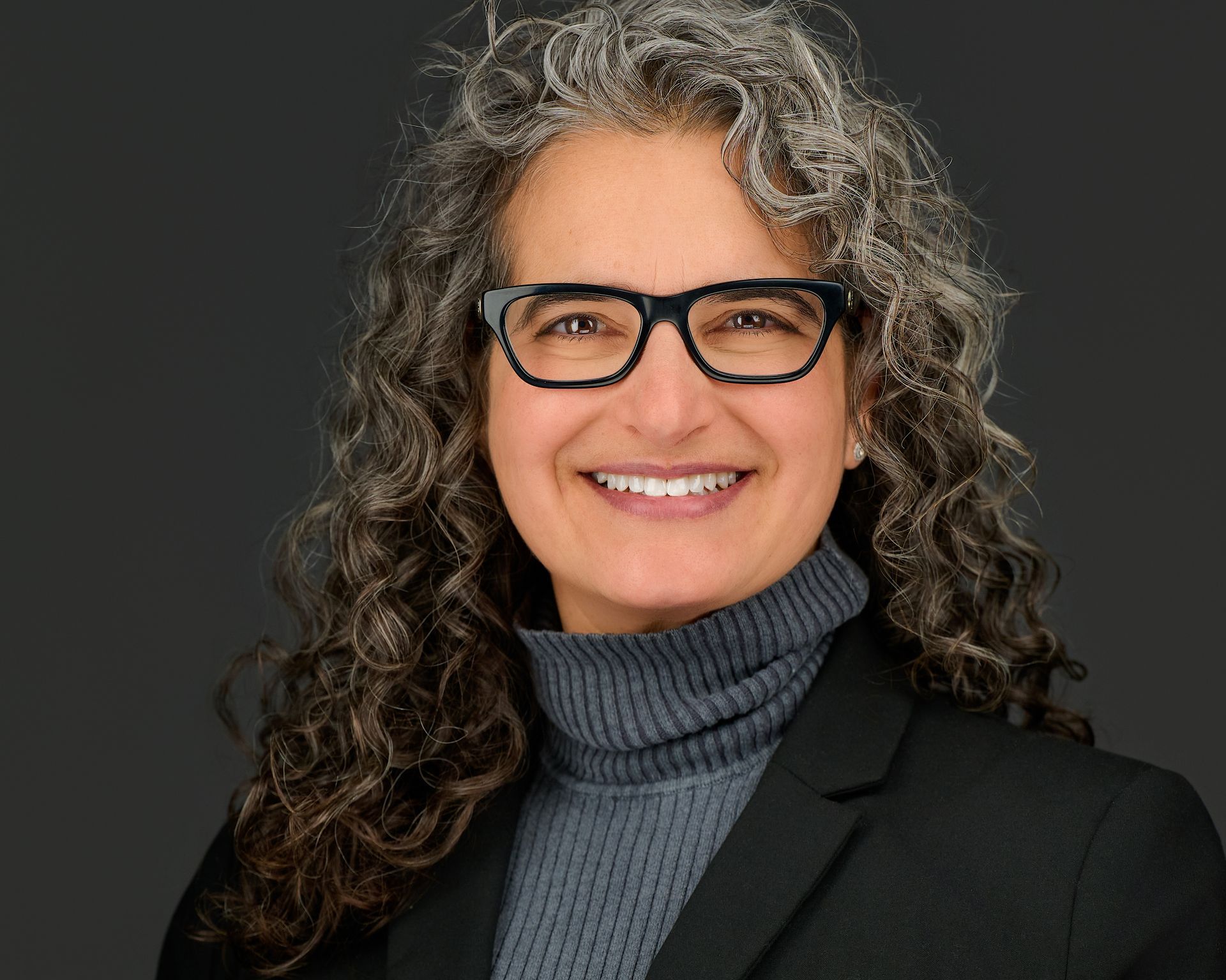A woman wearing glasses and a grey jacket is smiling.