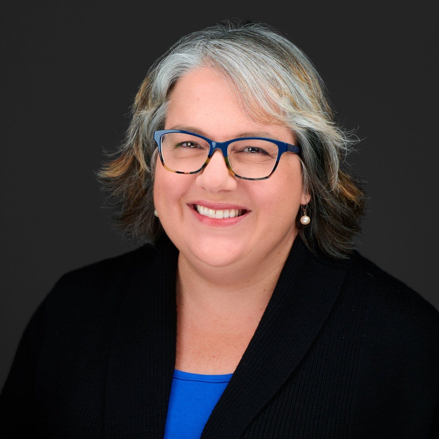 Woman with gray hair, glasses, smiling, wearing blue shirt and black jacket.