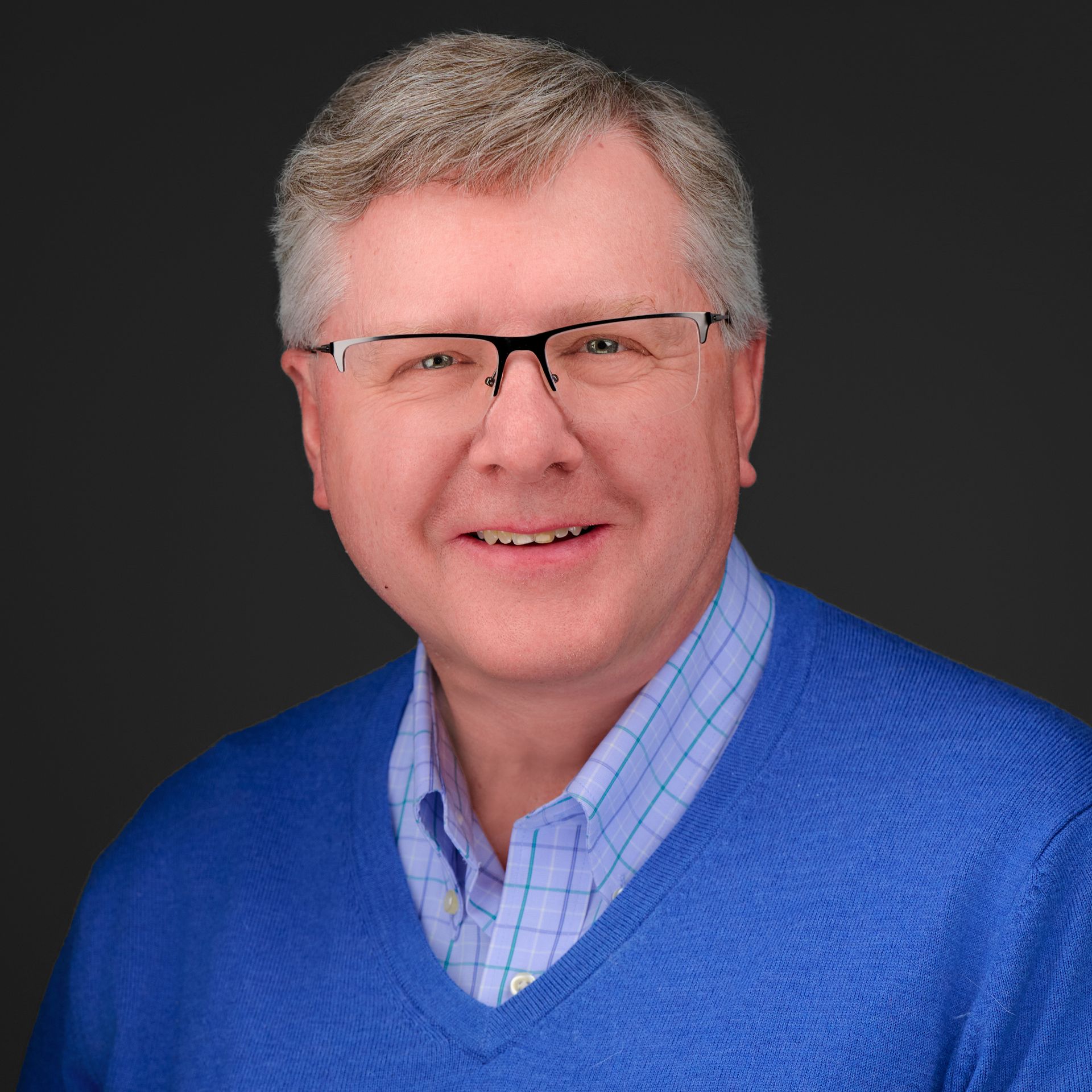 Man with glasses smiles, wearing blue sweater over light blue plaid shirt, against dark gray background.