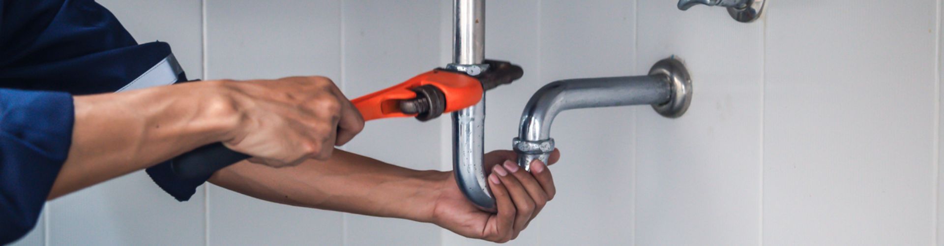 Plumber using an orange wrench to work on a silver pipe under a sink.