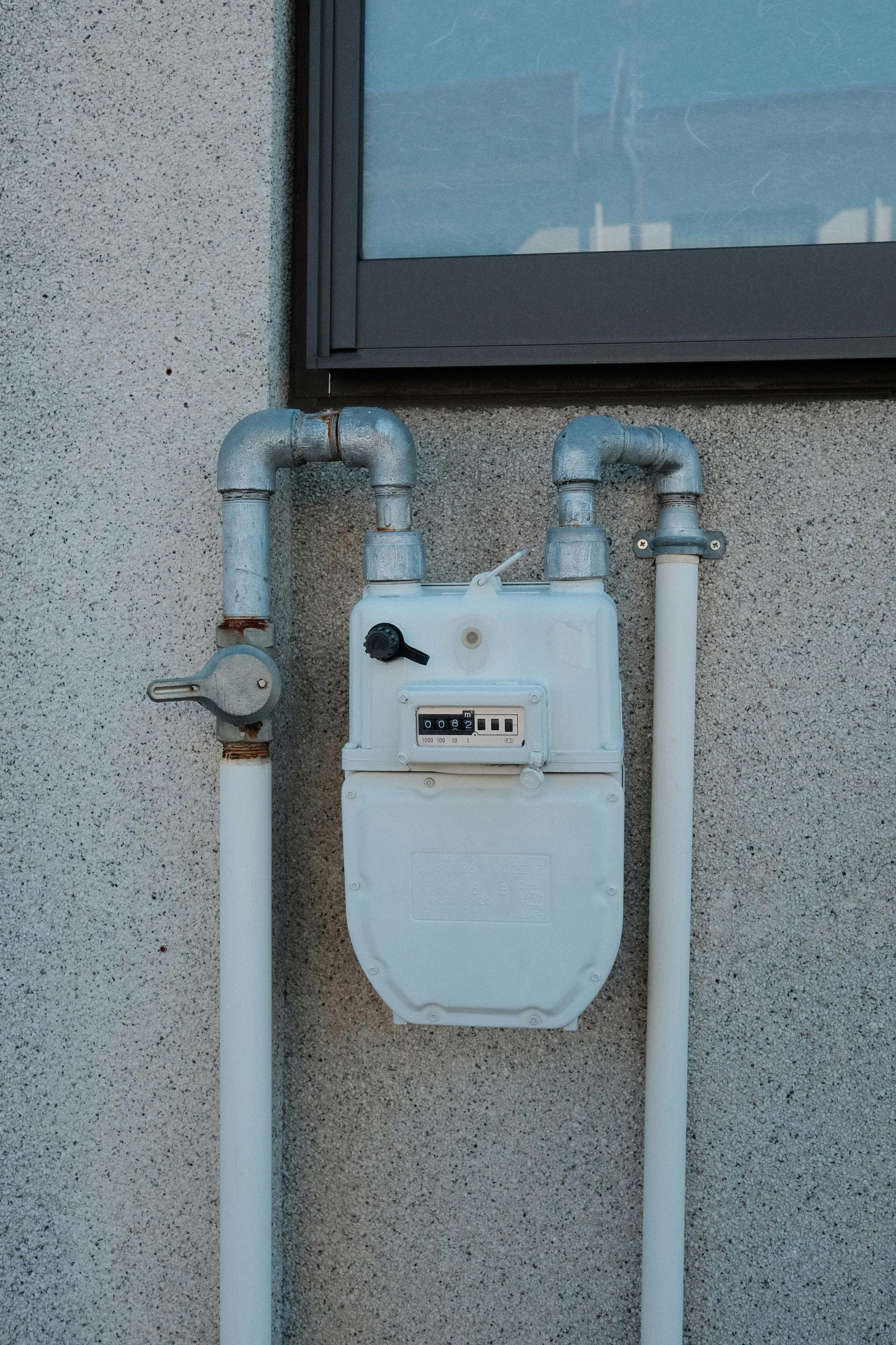 Gas meter and pipes mounted on an exterior stucco wall beneath a window.