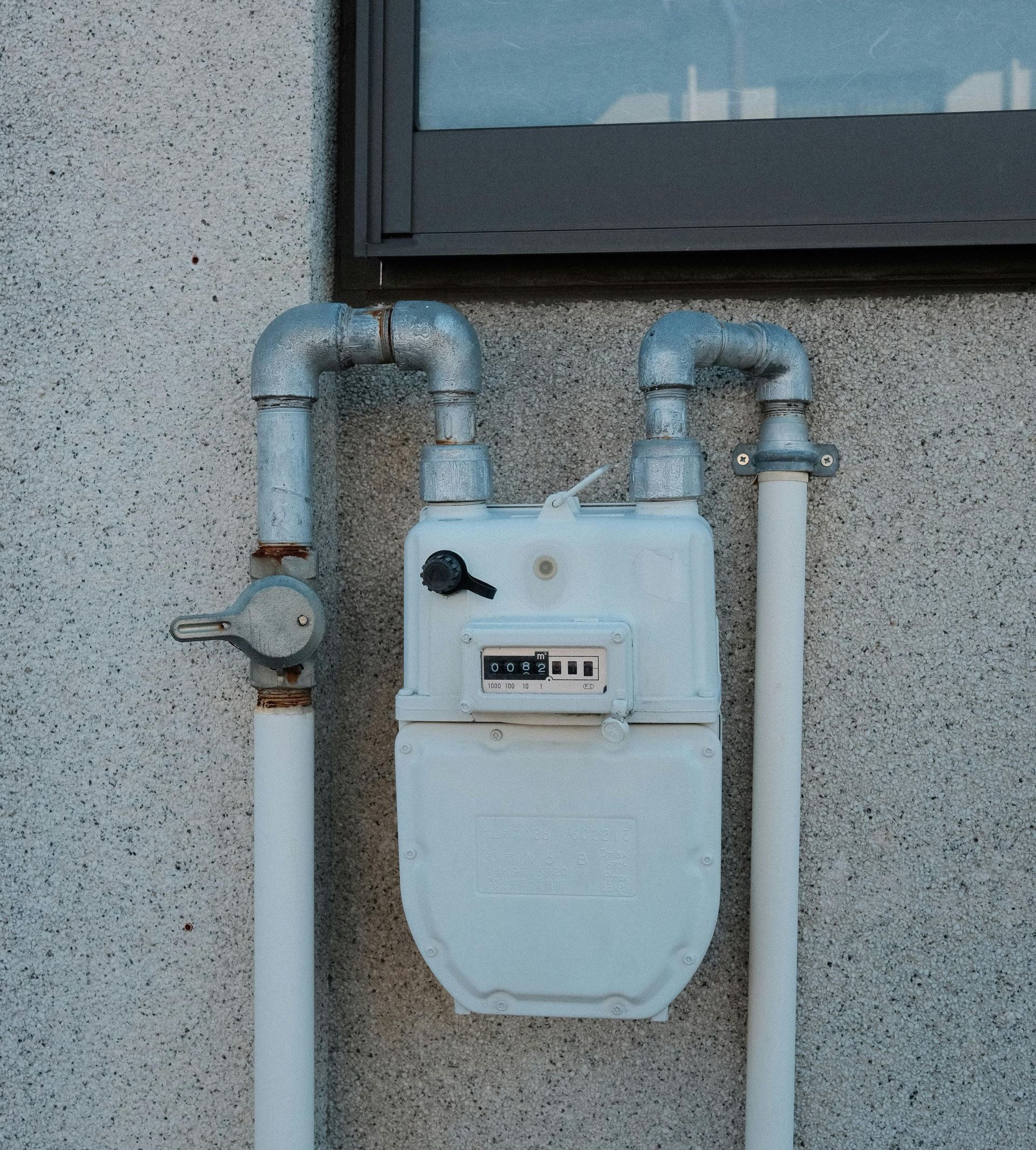Wall-mounted gas meter with silver pipes on a speckled exterior wall, beneath a window.