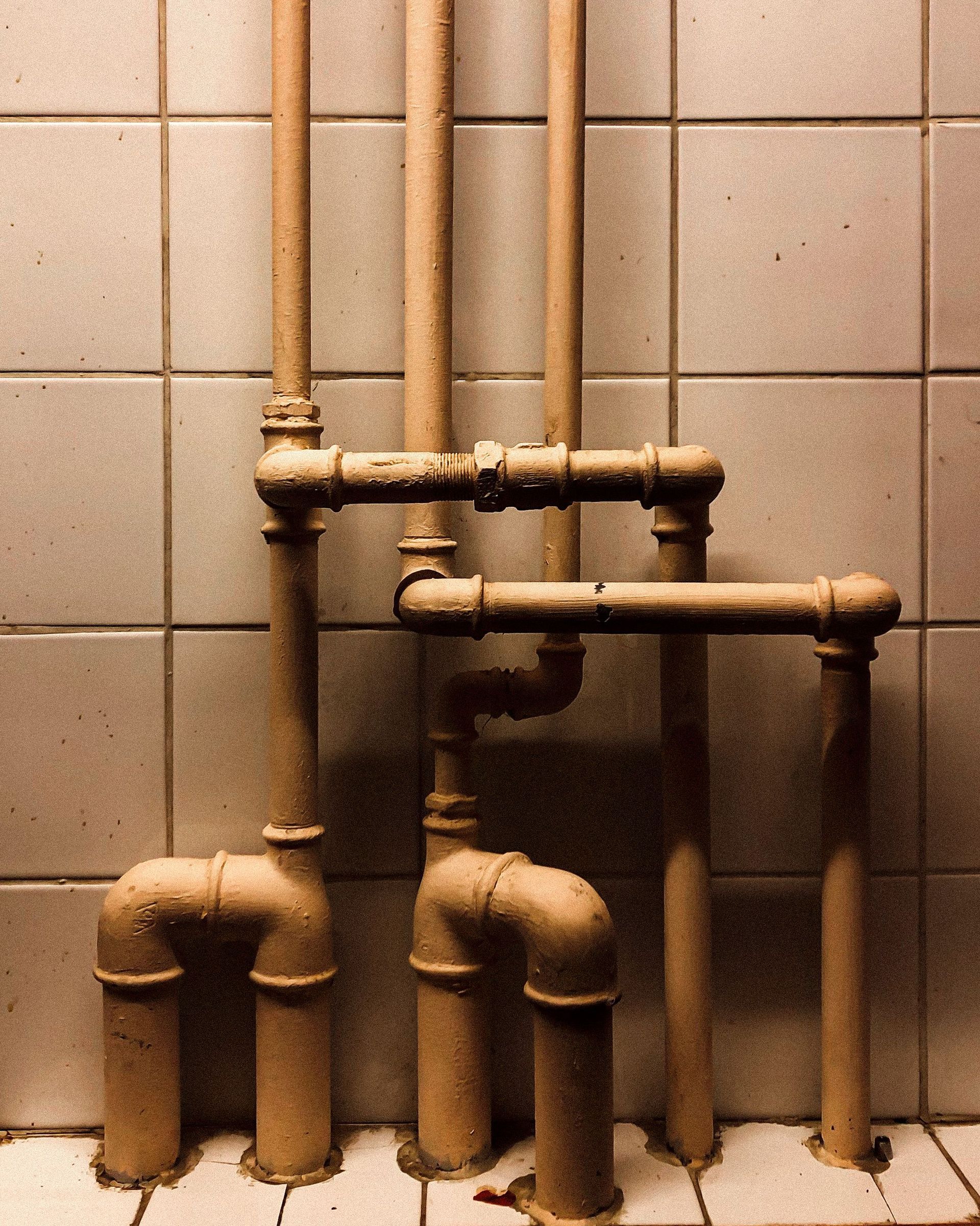 Rusty exposed pipes against a beige tiled wall in a bathroom or utility room.