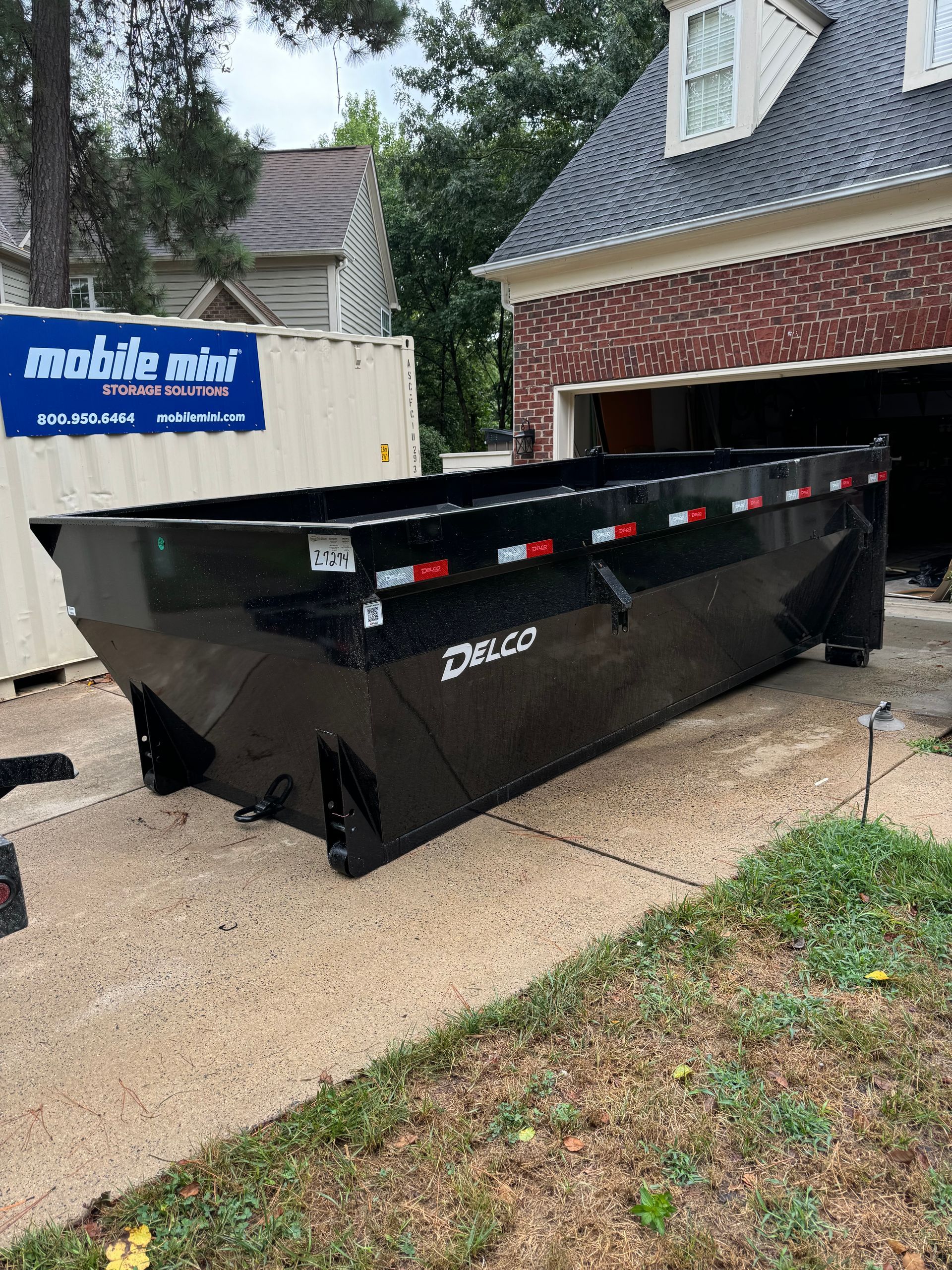 A large black dumpster is parked in front of a brick house.
