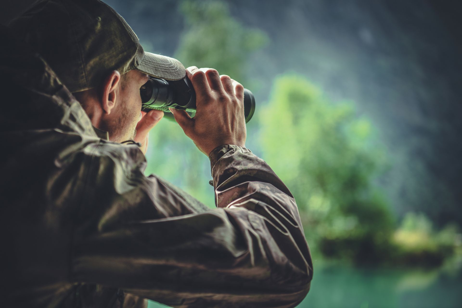 Person in a cap using binoculars in a misty outdoor landscape, looking toward the water
