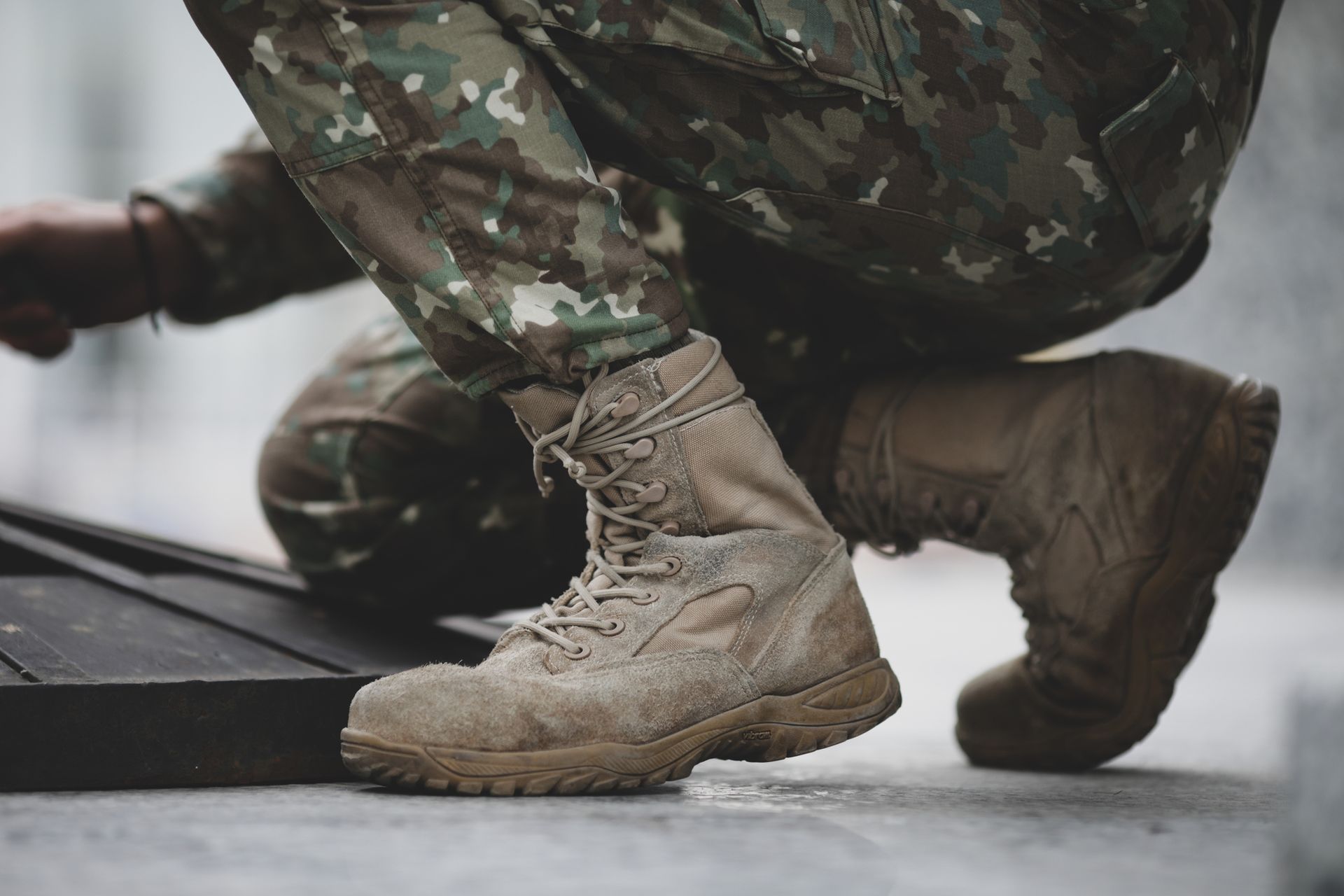 Camouflage-clad soldier’s tan combat boots crouching on a wet surface