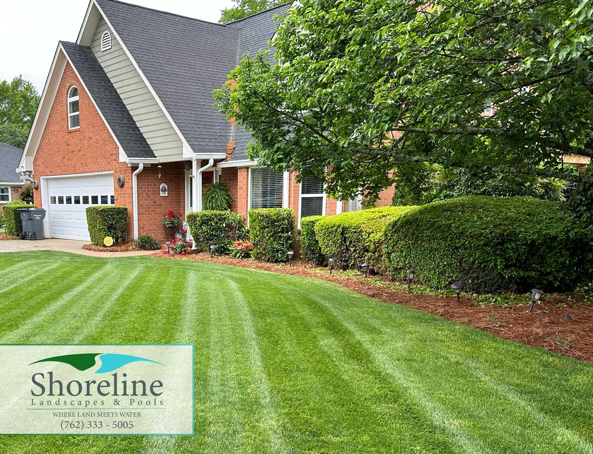 Lush green lawn with striped mowing pattern in front of a brick house with manicured hedges and Shoreline Landscaping logo.