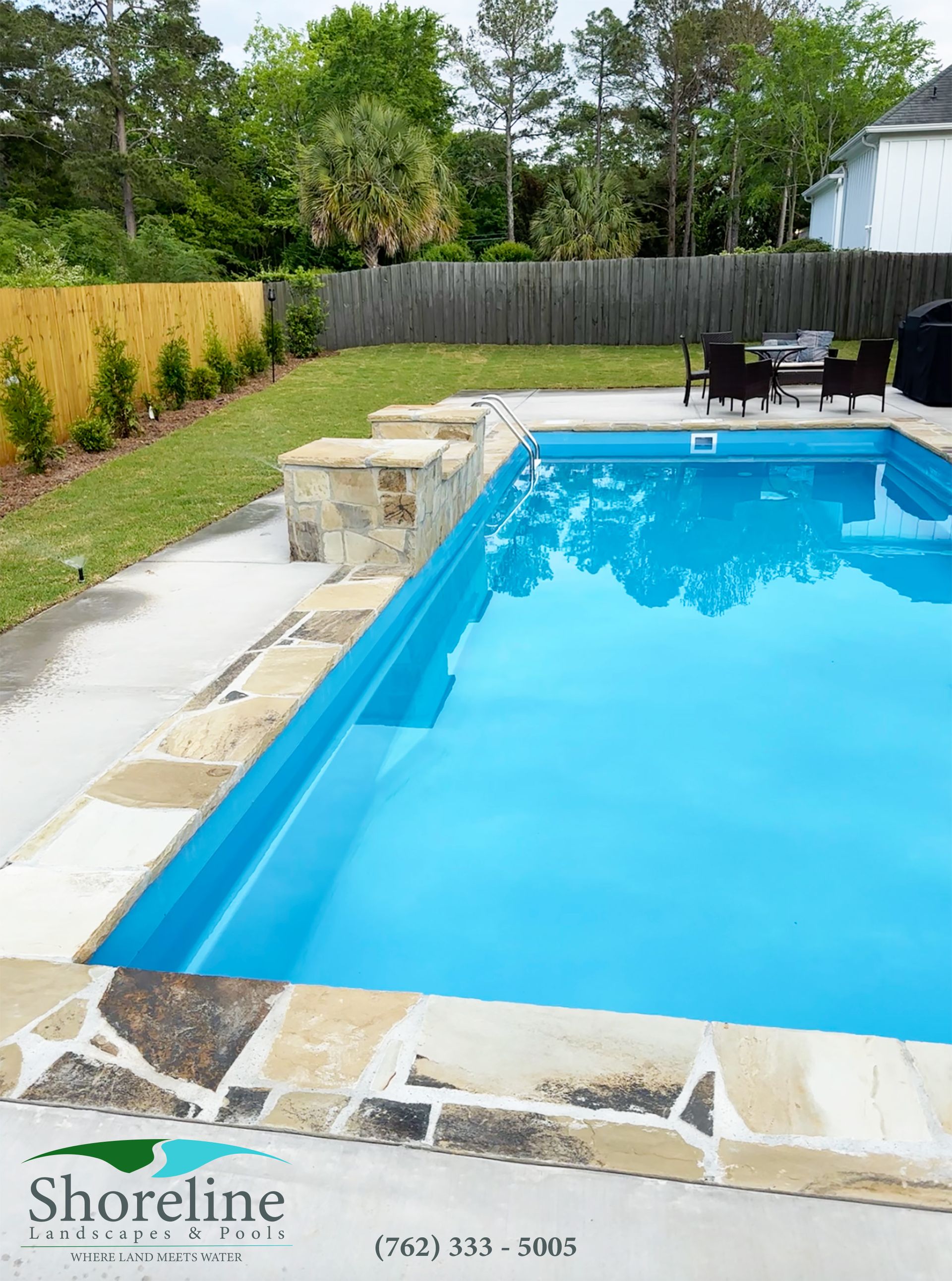 Pool with blue water and stone coping. Lawn, fence, and seating area are in the background.