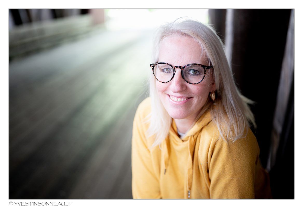 Une femme portant des lunettes et un sweat à capuche jaune sourit à la caméra