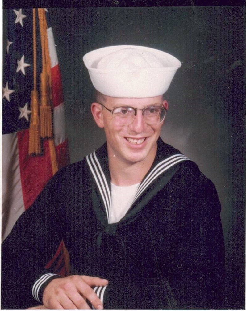 A man in a navy uniform is smiling in front of an american flag