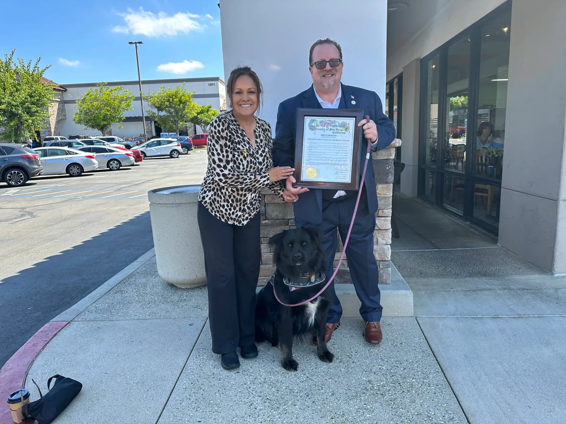 A man and a woman are standing next to a black dog holding a certificate.