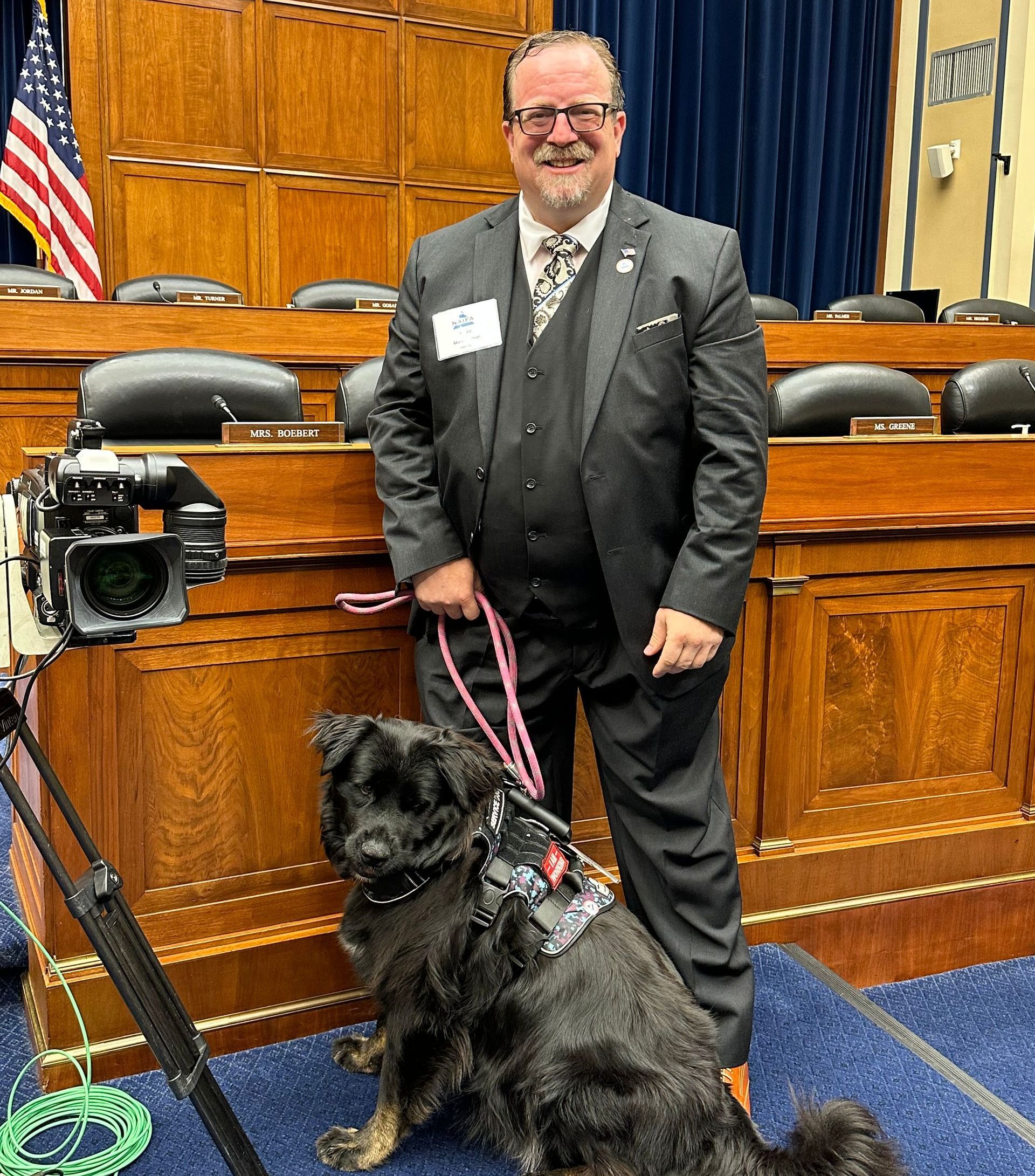 A man in a suit is standing next to a dog on a leash in front of a camera.