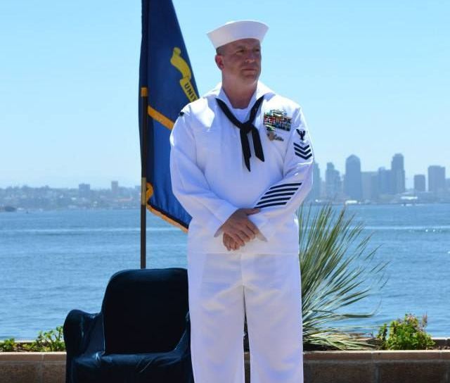 A man in a navy uniform stands in front of a flag