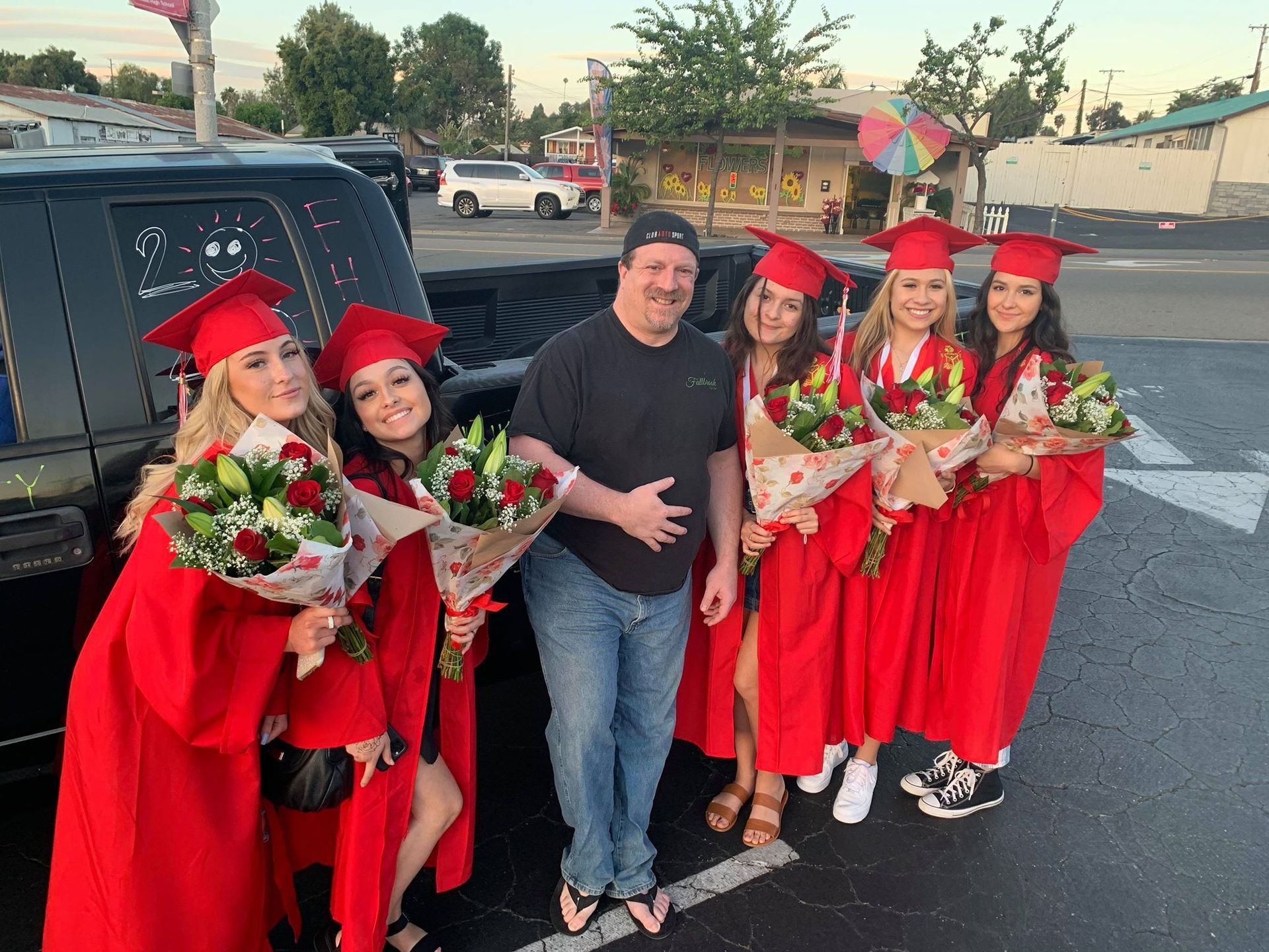 A group of graduates are posing for a picture with a man in a parking lot.