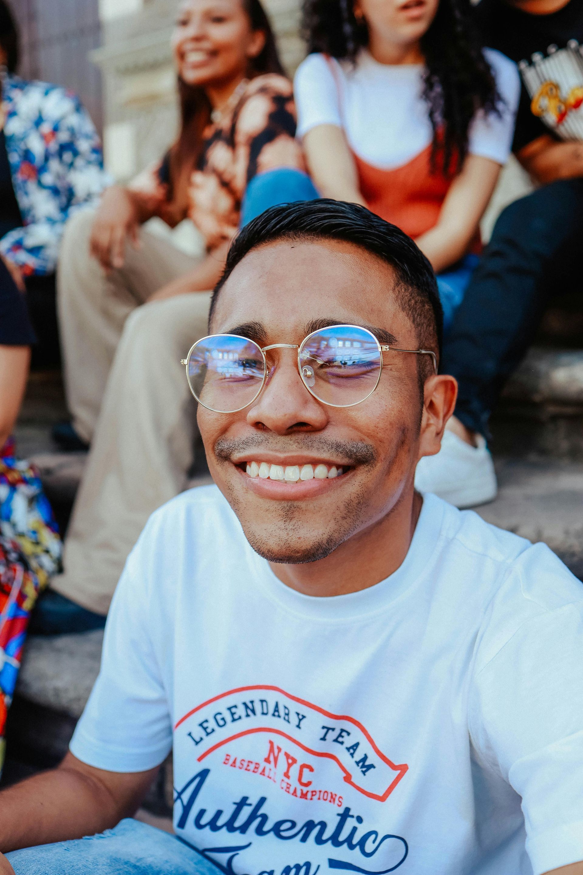 Smiling person in glasses and white t-shirt, sitting on steps with others.