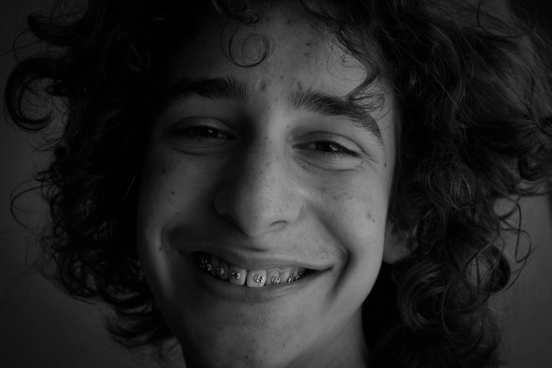 A young man with curly hair and braces is smiling in a black and white photo.