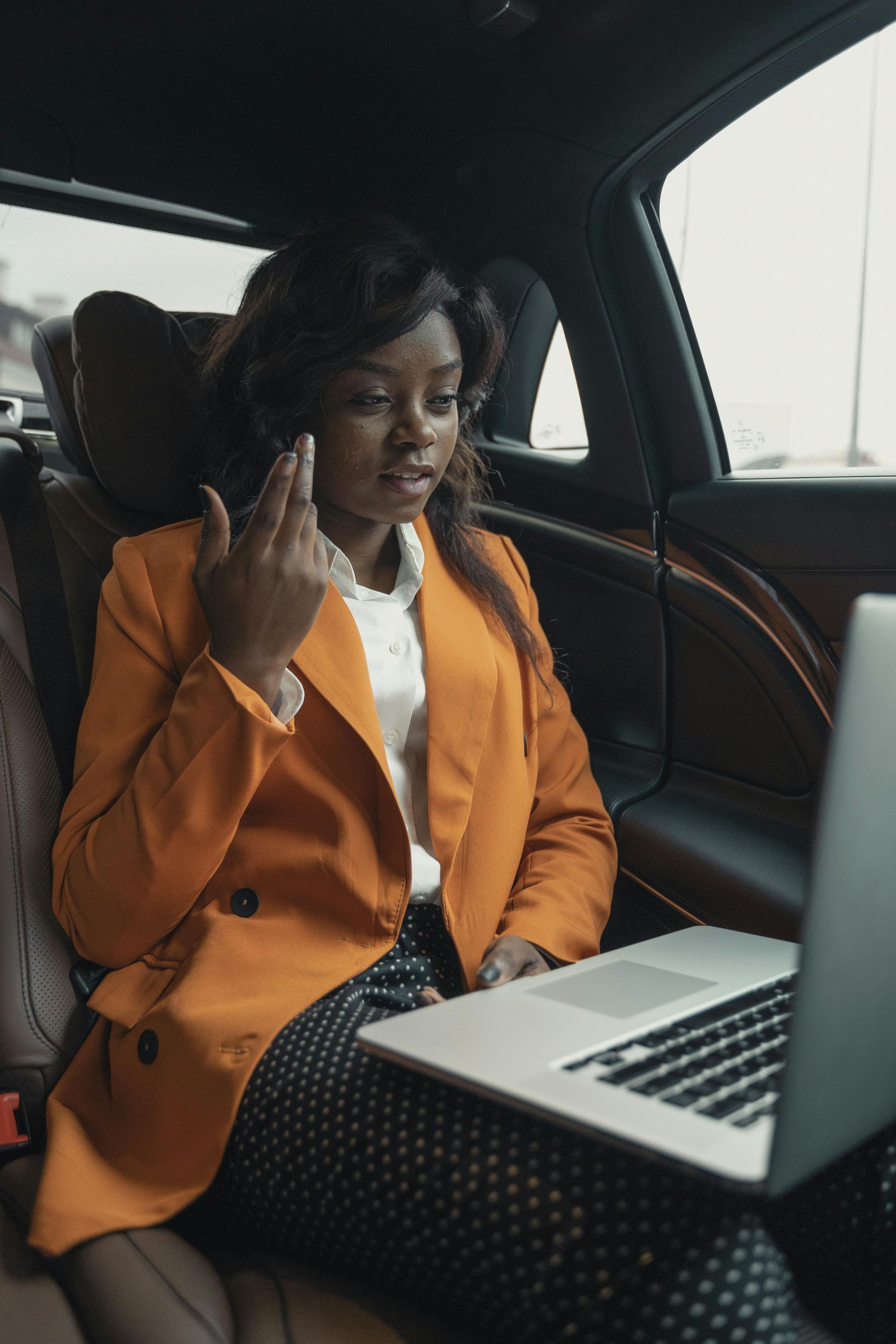 Woman in orange blazer on a video call in a car, looking at a laptop, gesturing with her fingers.