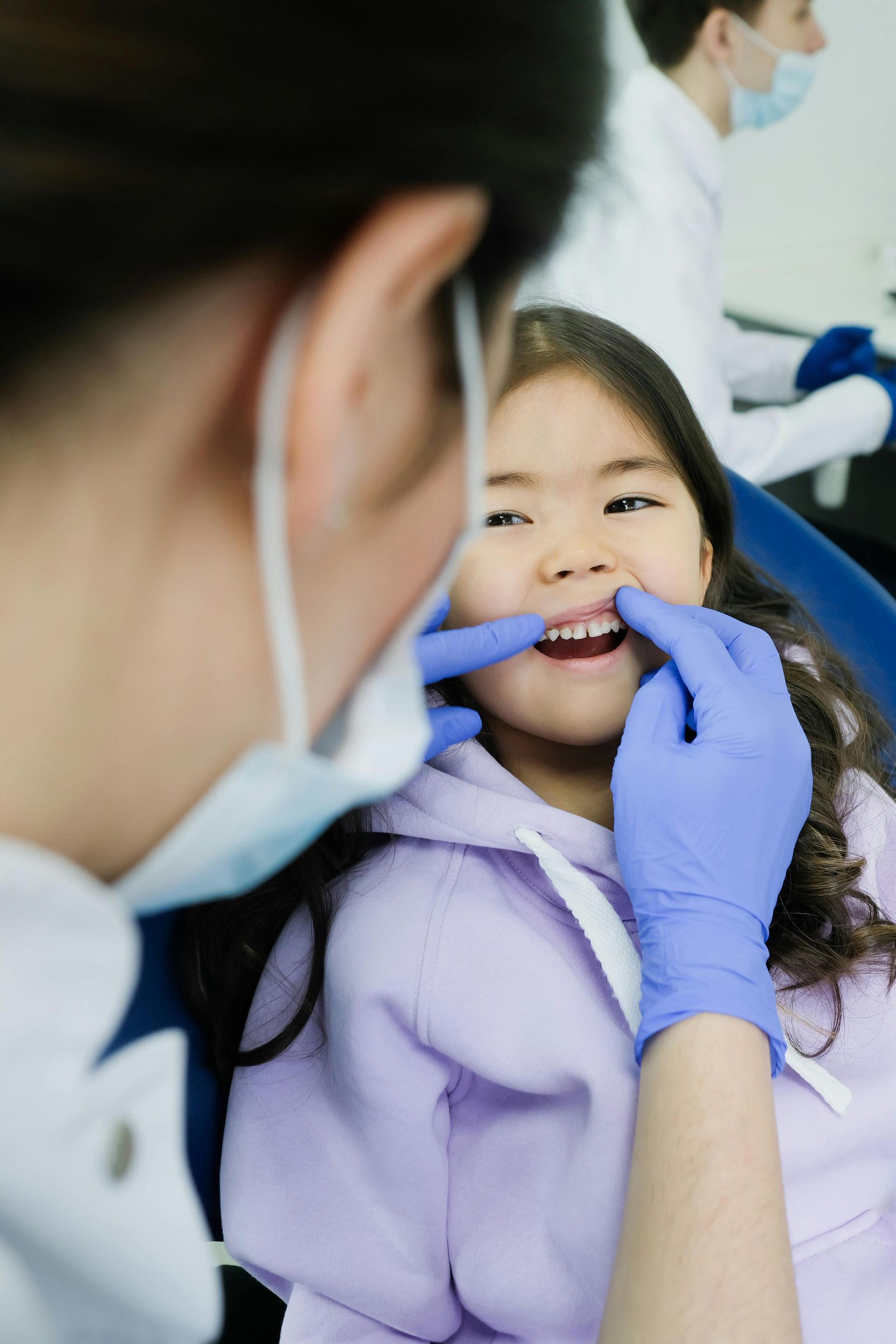 Dentist examining a child's teeth. Child smiles, wearing purple hoodie. Dentist wears mask, gloves in office.