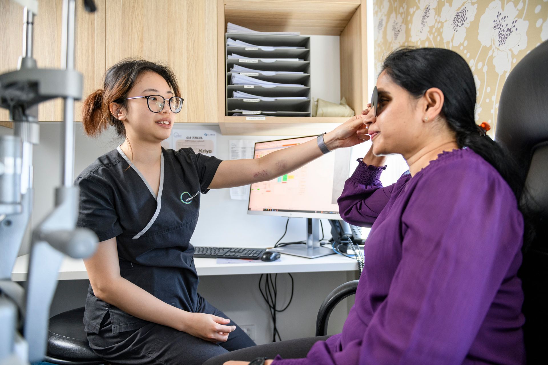 An eye examination machine, white and gray, with attached monitor, on a desk with a laptop and a chair.