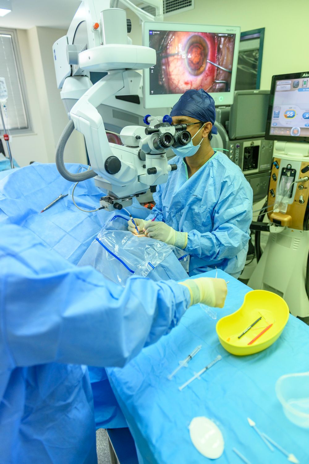 Surgeon performing eye surgery, using a microscope. Patient draped in blue, surgical tools on the table.