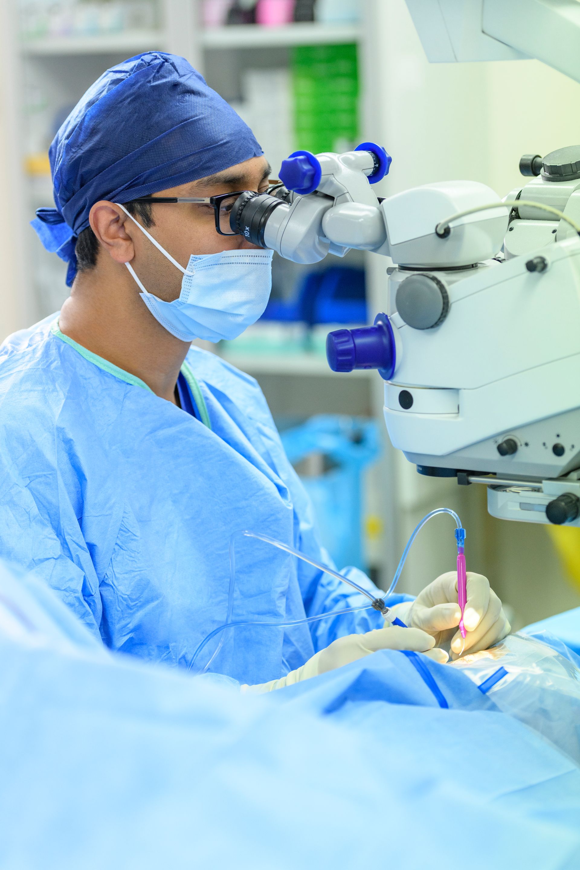 Surgeon in operating room using microscope, performing surgery, wearing blue scrubs and mask.