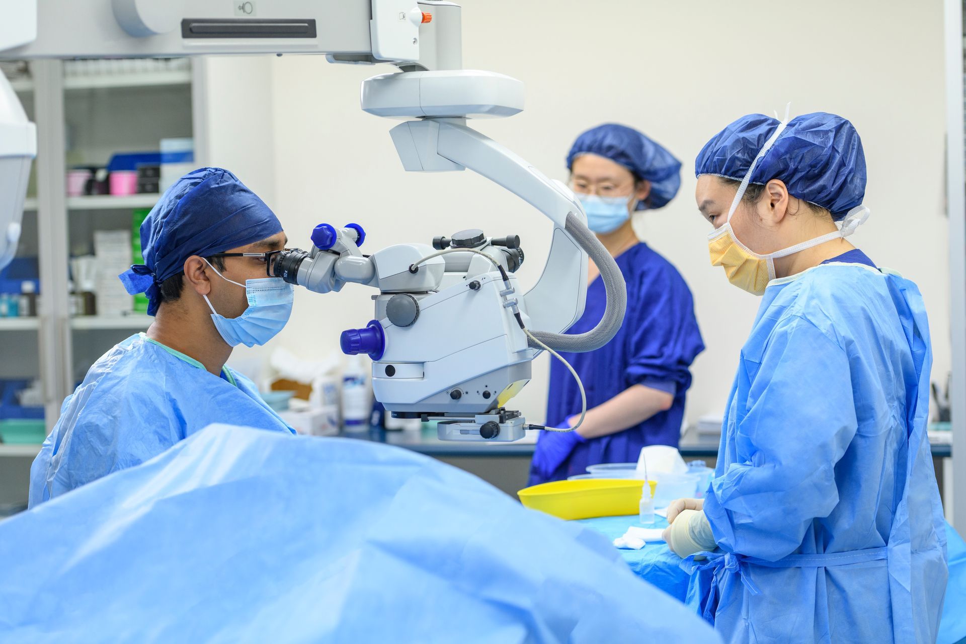 Eye surgery being performed in a sterile operating room with a surgical microscope and masked medical staff.
