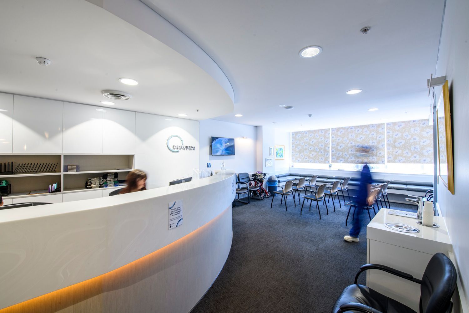 Reception area in a modern medical clinic; white walls, curved desk, waiting chairs, person in blue scrubs.