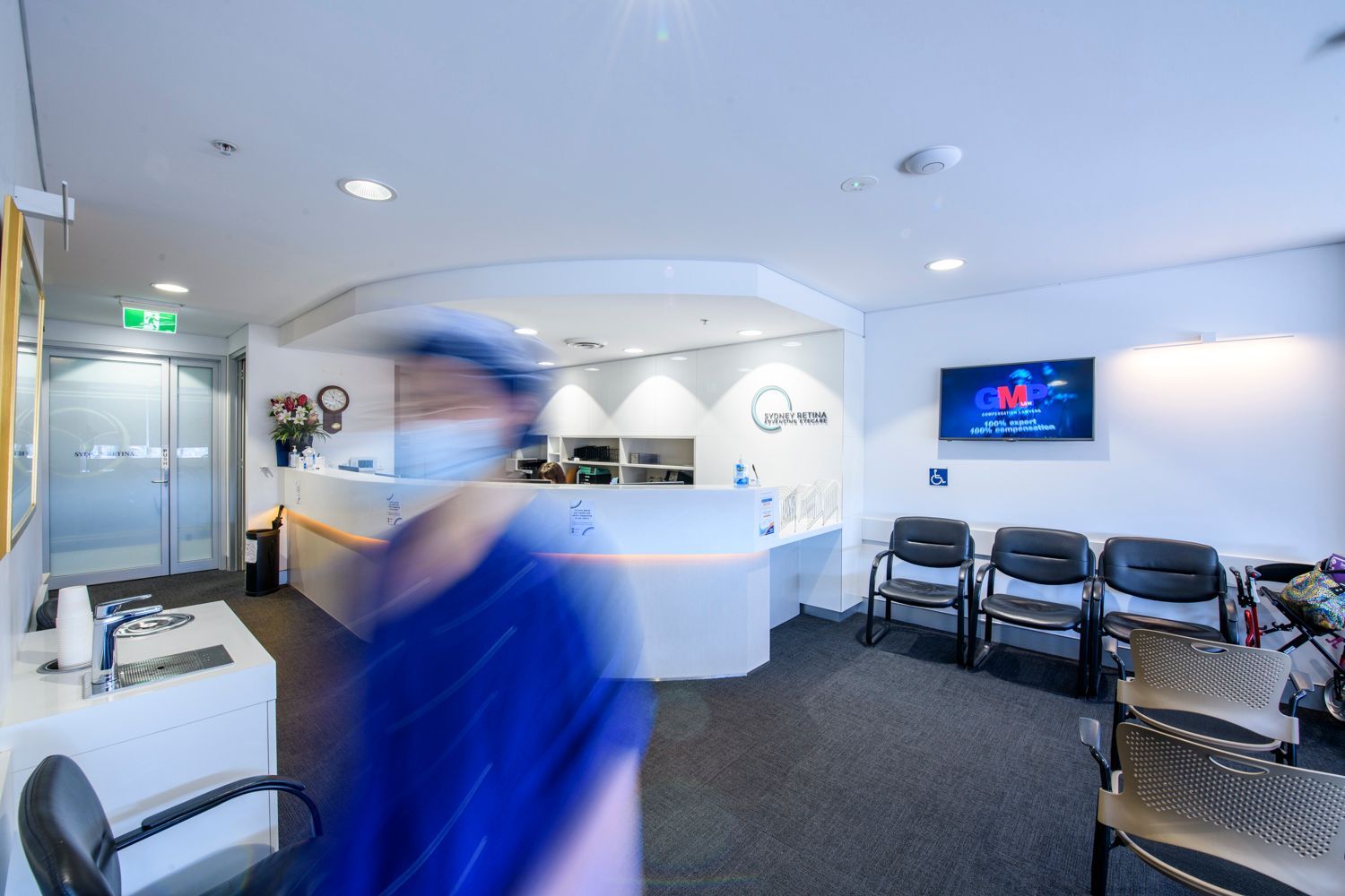 Reception area with a blurred person in blue scrubs, reception desk, and waiting chairs.