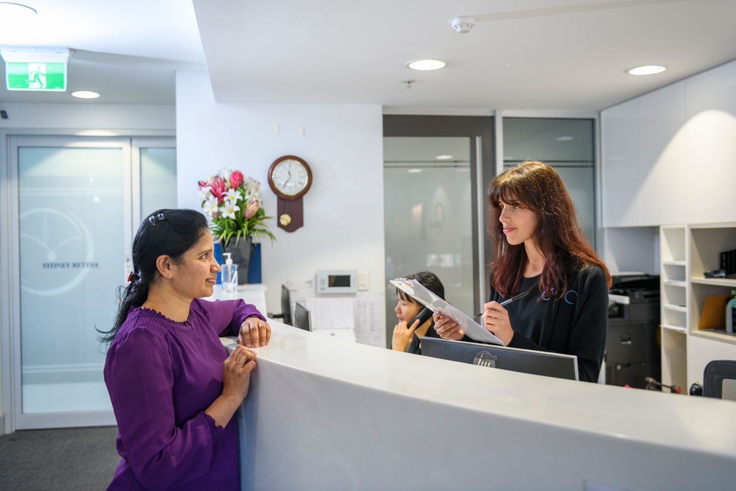 Man at a reception desk, talking to a receptionist. Modern, white office with chairs visible in the background.