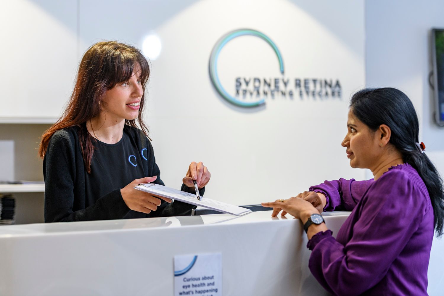 Woman at a clinic reception desk assisting another woman. Clinic sign visible.