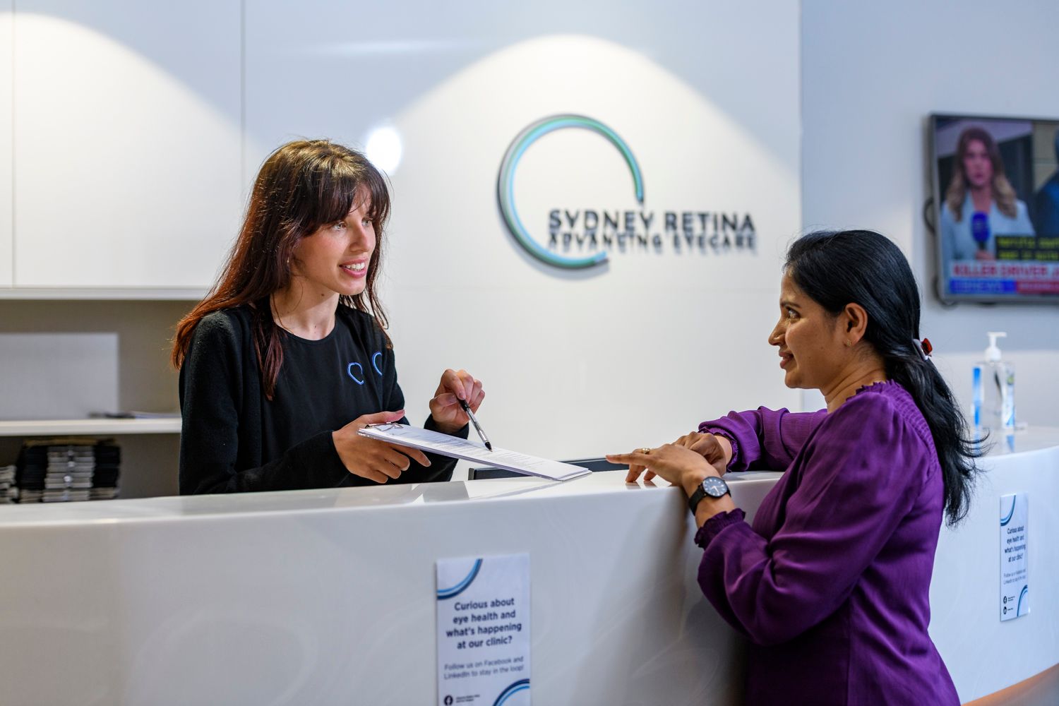 Woman at a reception desk assisting a patient. Sign above reads 