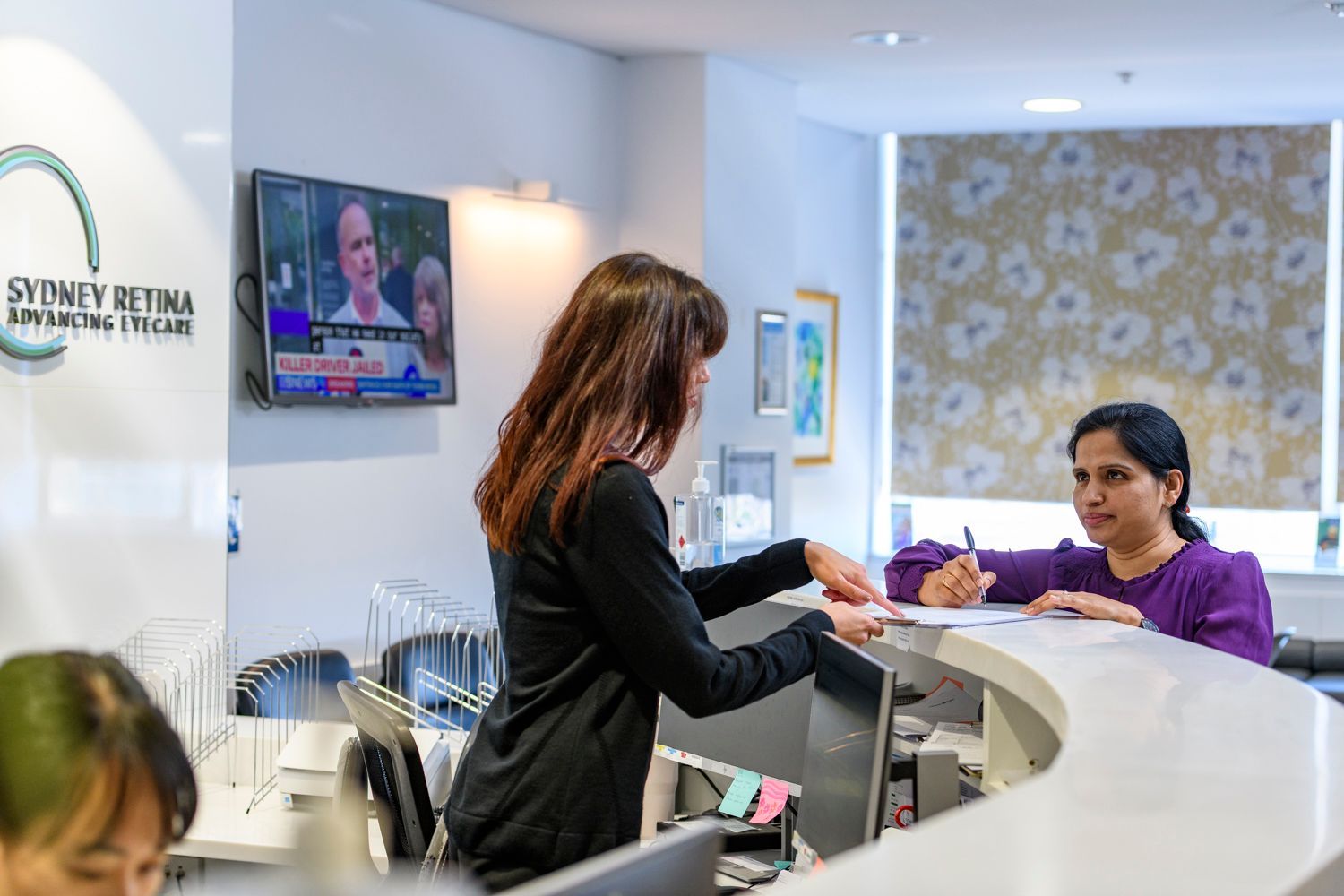 Receptionist assisting a patient at Sydney Retina clinic. White desk, TV playing, light colors.
