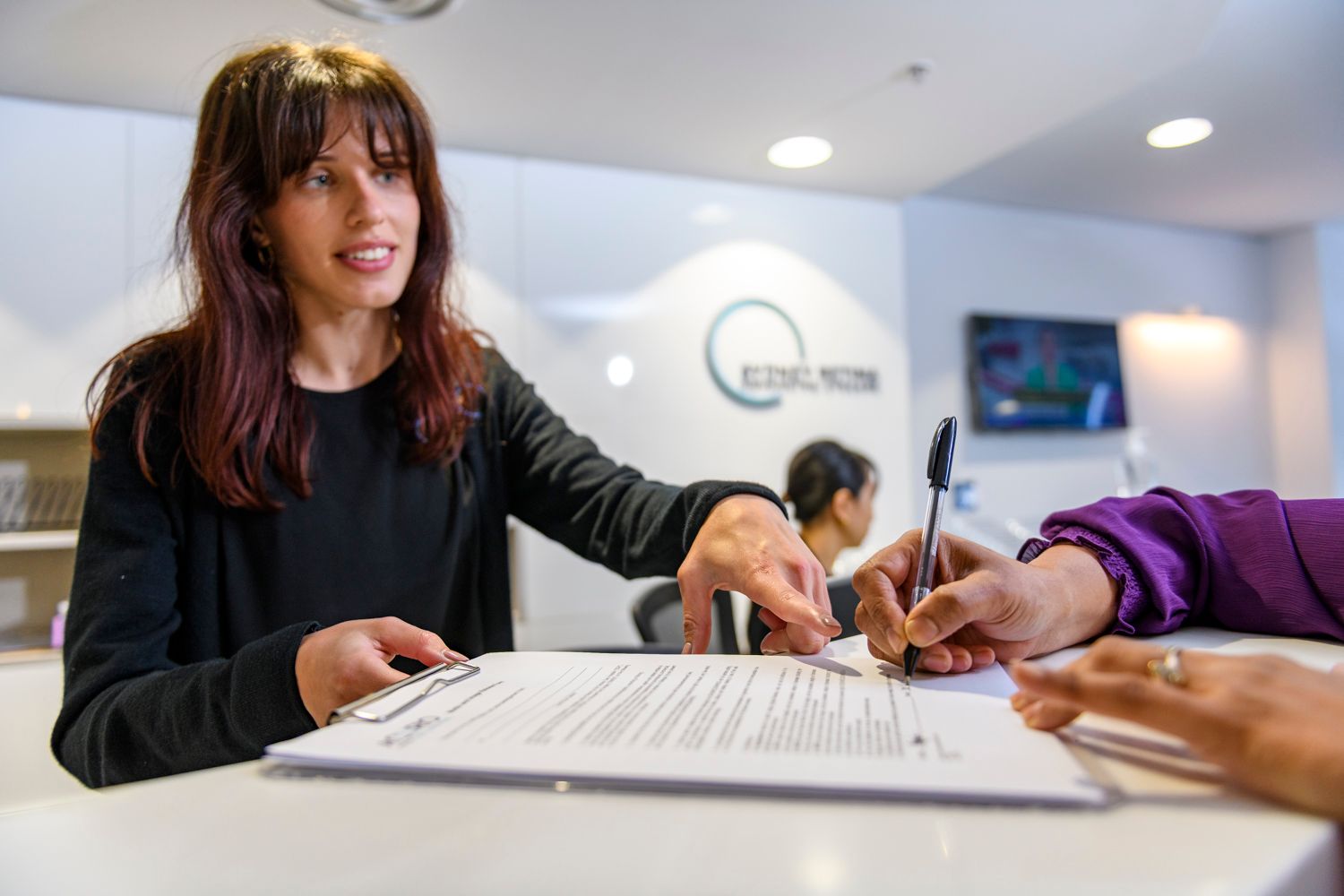 Woman at a reception desk assisting a person signing paperwork. Modern office setting.