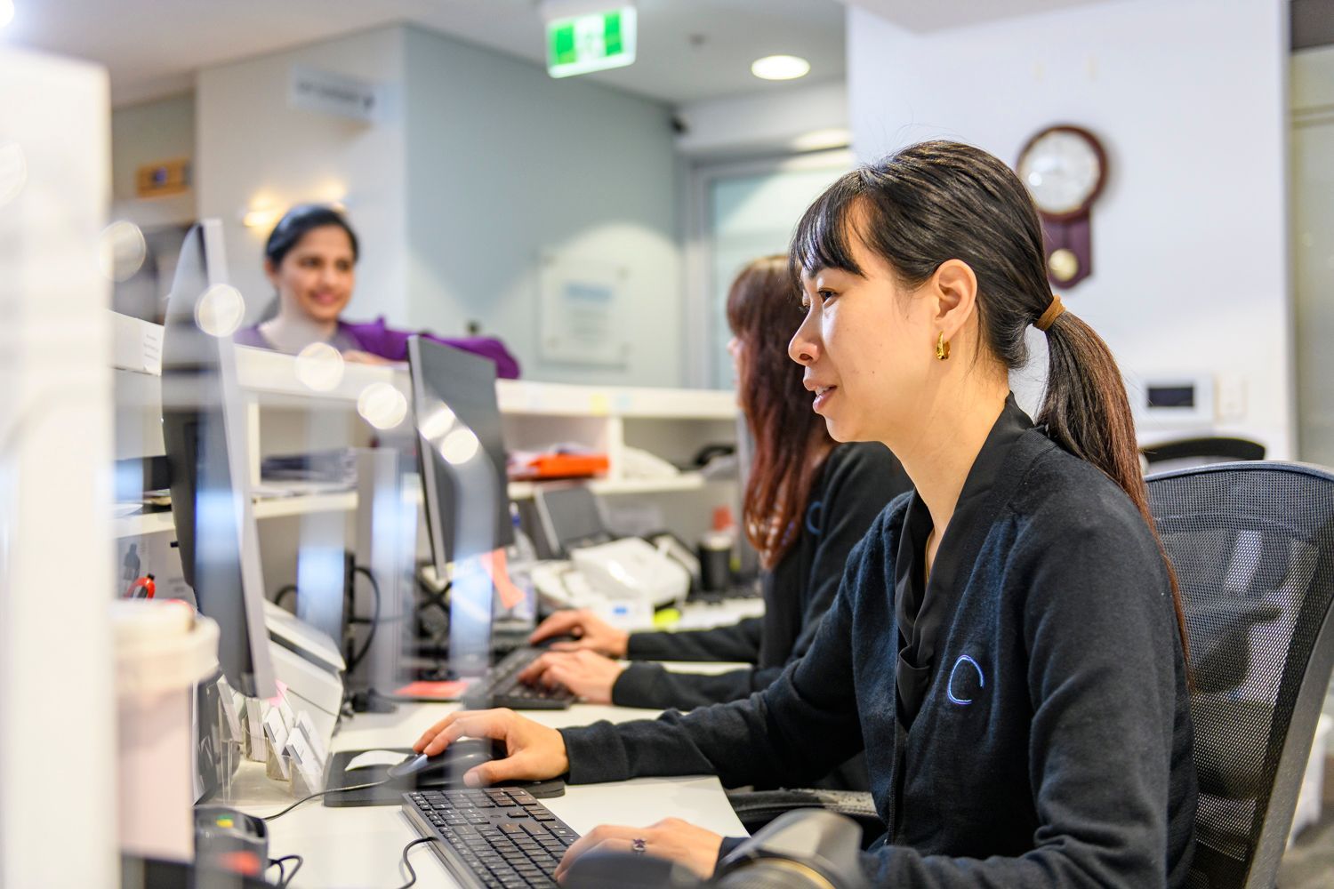 Three office workers at computer desks, typing and looking at monitors; one smiles at the others.