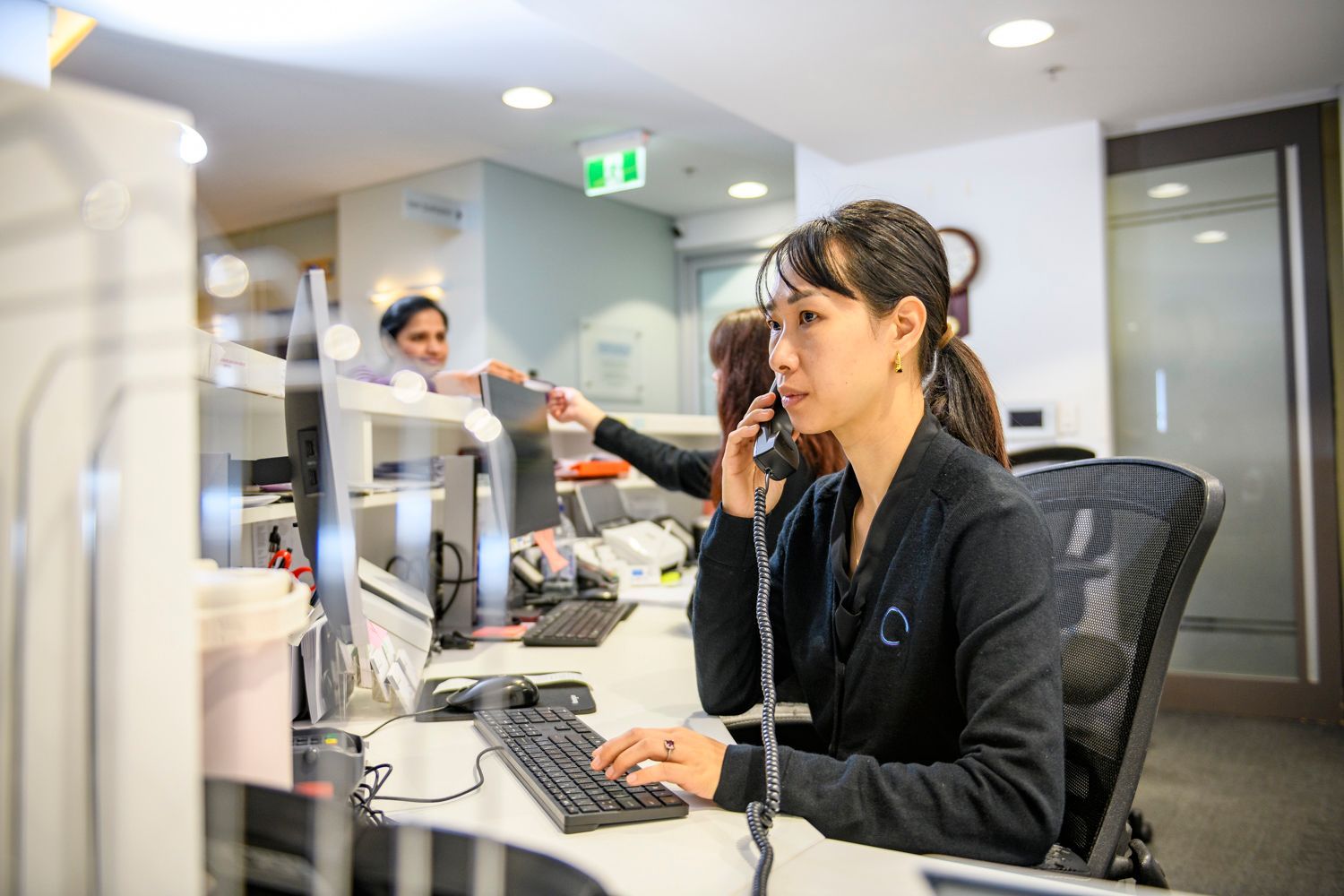 Woman in office, on phone, typing at a computer. Other people and cubicles are in the background.