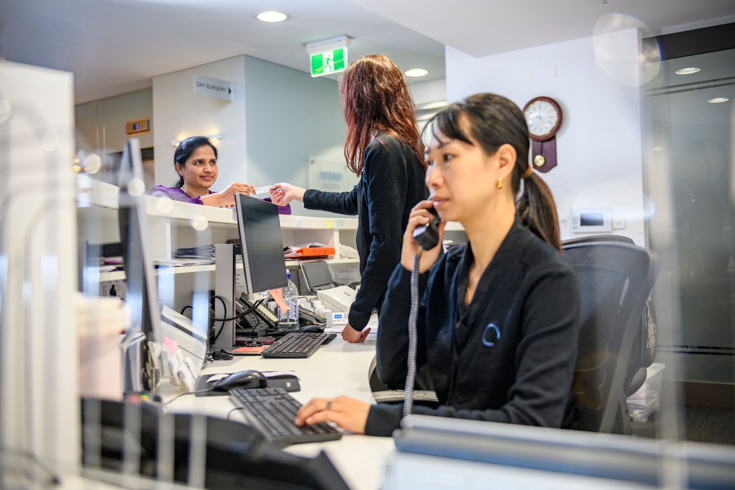 Medical office reception desk with staff: one on phone, two assisting patients.