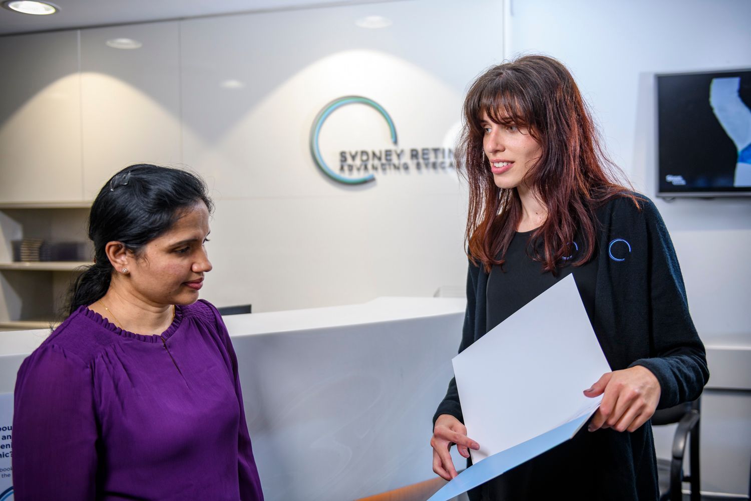 Woman in purple shirt at a reception desk; staffer in black jacket holds papers and gestures. Sydney Retina Clinic sign.