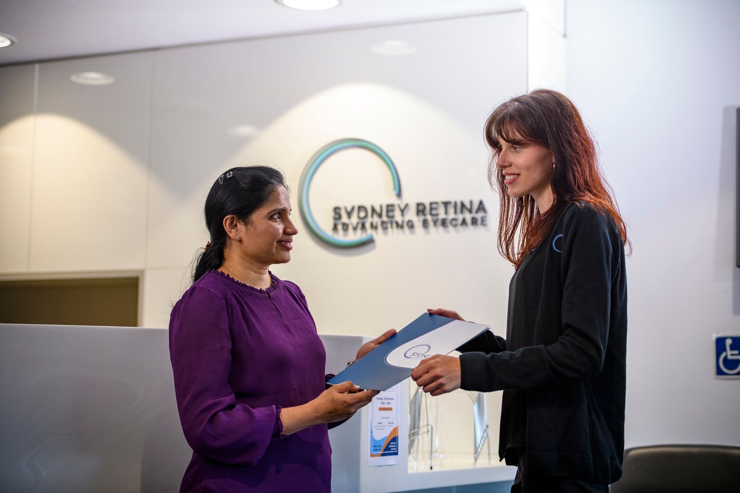 Woman hands file to another woman at the Sydney Retina Eye Care reception.