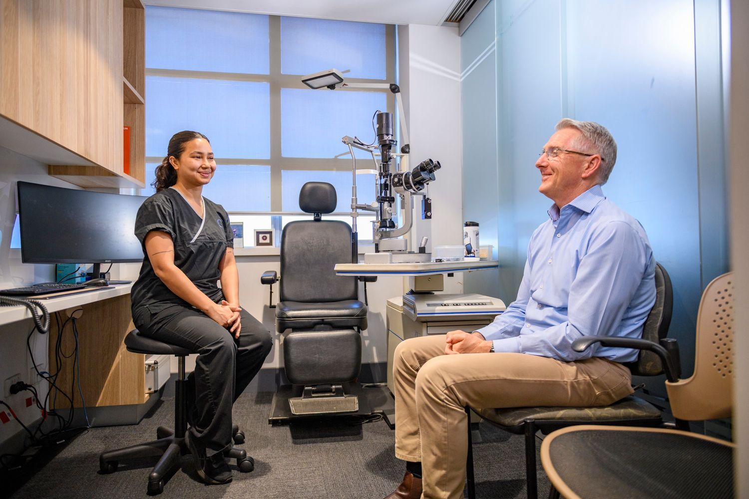 Optometrist and patient in an exam room. Doctor smiles at patient sitting, equipment behind.