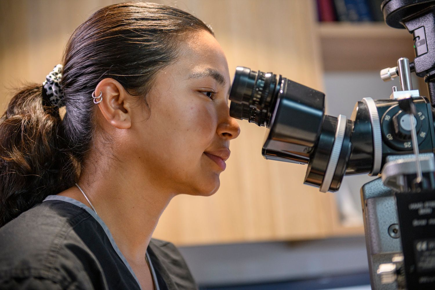 Woman looking into a black ophthalmic microscope in a clinical setting.