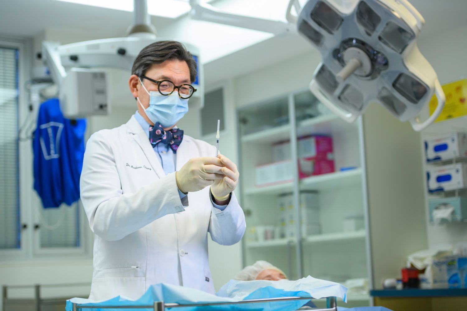 Surgeon in operating room prepares injection. Surgical light, patient, and medical equipment in the background.
