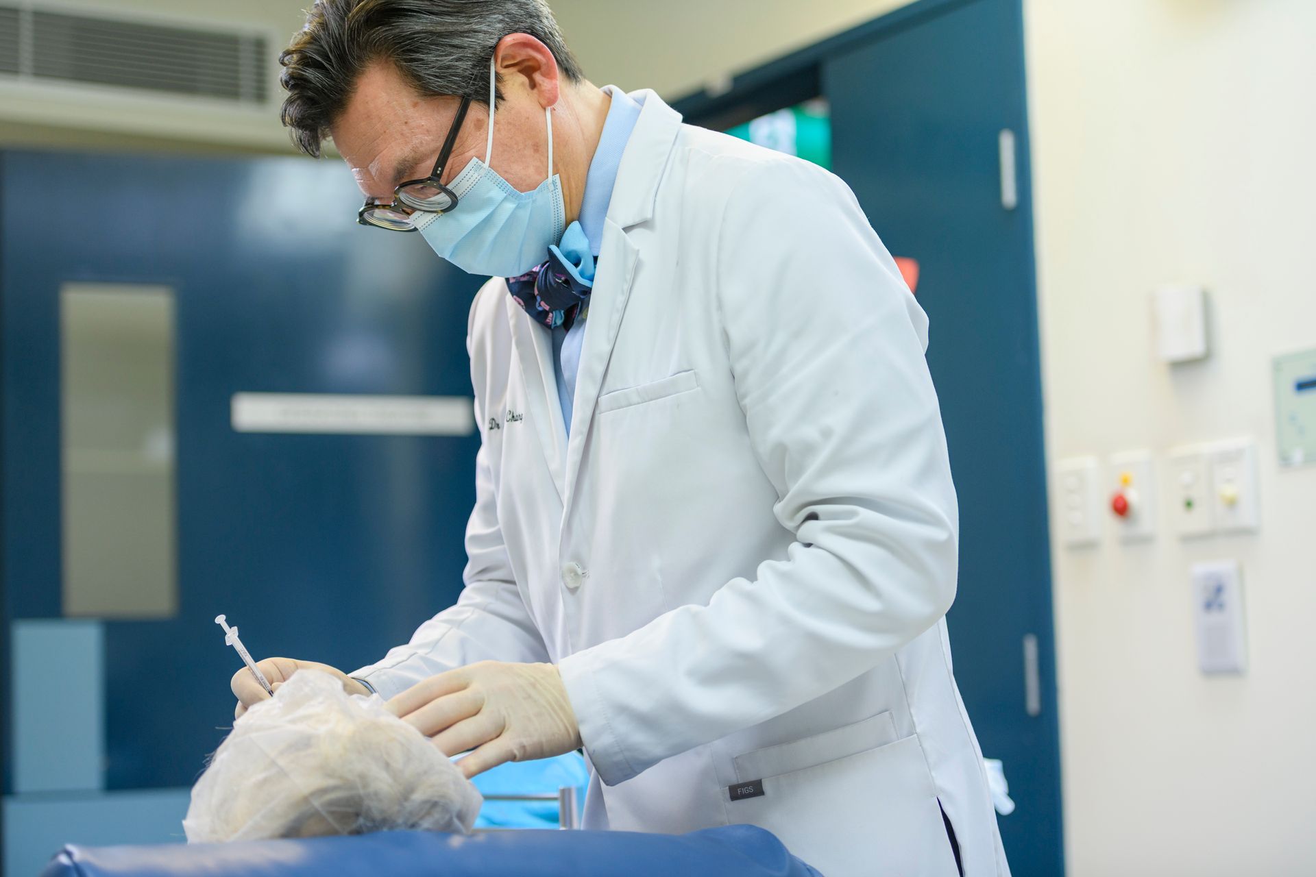 Doctor in lab coat and mask administering an injection to a fluffy, white object in a clinical setting.