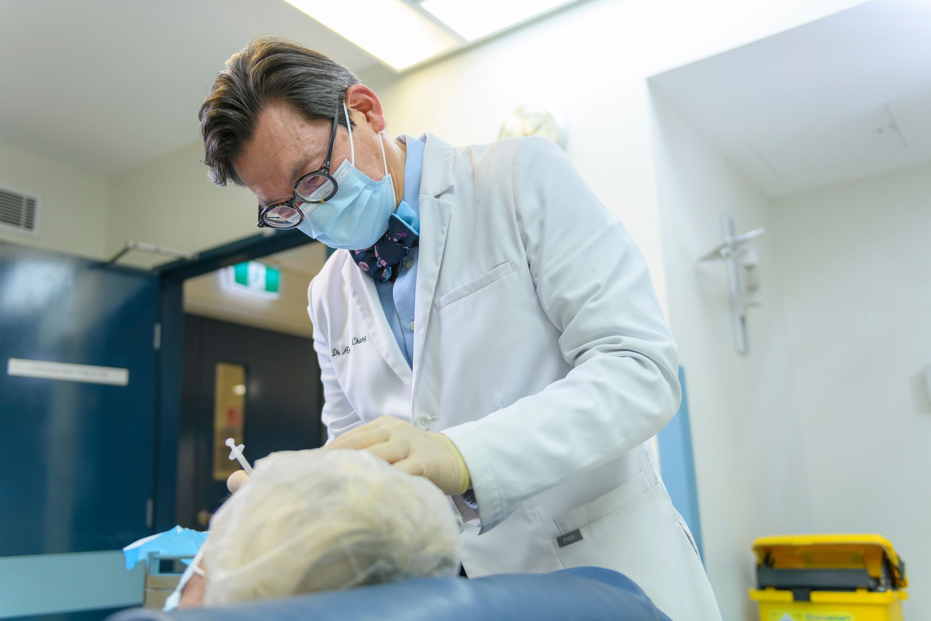 Doctor in mask, gloves, and lab coat, administering treatment to a patient in a medical setting.