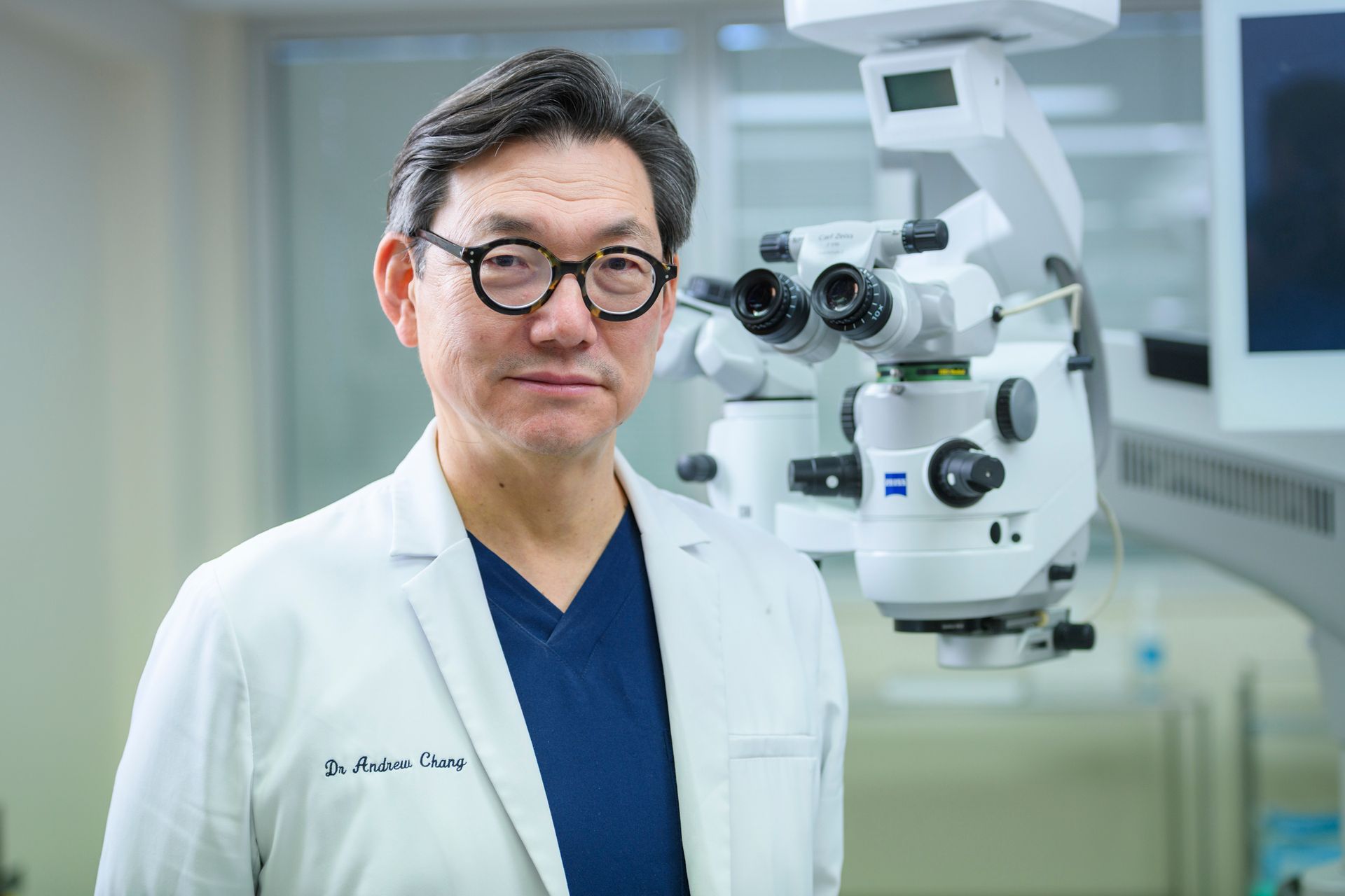 Man in doctor's coat stands near surgical microscope in medical setting. He wears glasses and smiles slightly.